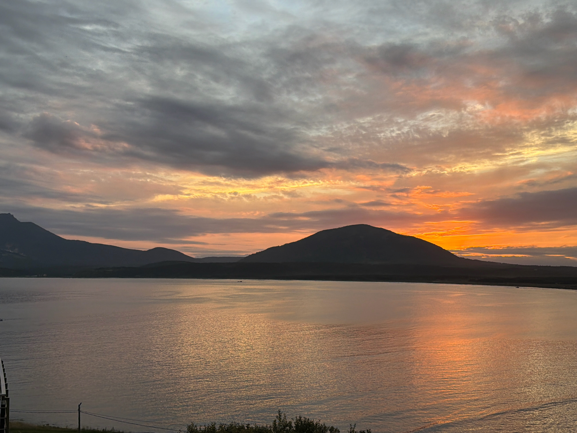 The image depicts a sunset over a body of water, with silhouetted mountains in the background. The sky is filled with clouds displaying hues of orange, pink, and gray. The water reflects the warm colors of the sunset. In the foreground, there is a glimpse of land with some vegetation and what appears to be a utility pole. The scene is likely a natural landscape, potentially a bay or a large lake, located near Yuzhno-Kurilsk, Russia, given the mountainous terrain and coastal proximity implied by the name. No people or specific activities are visible.