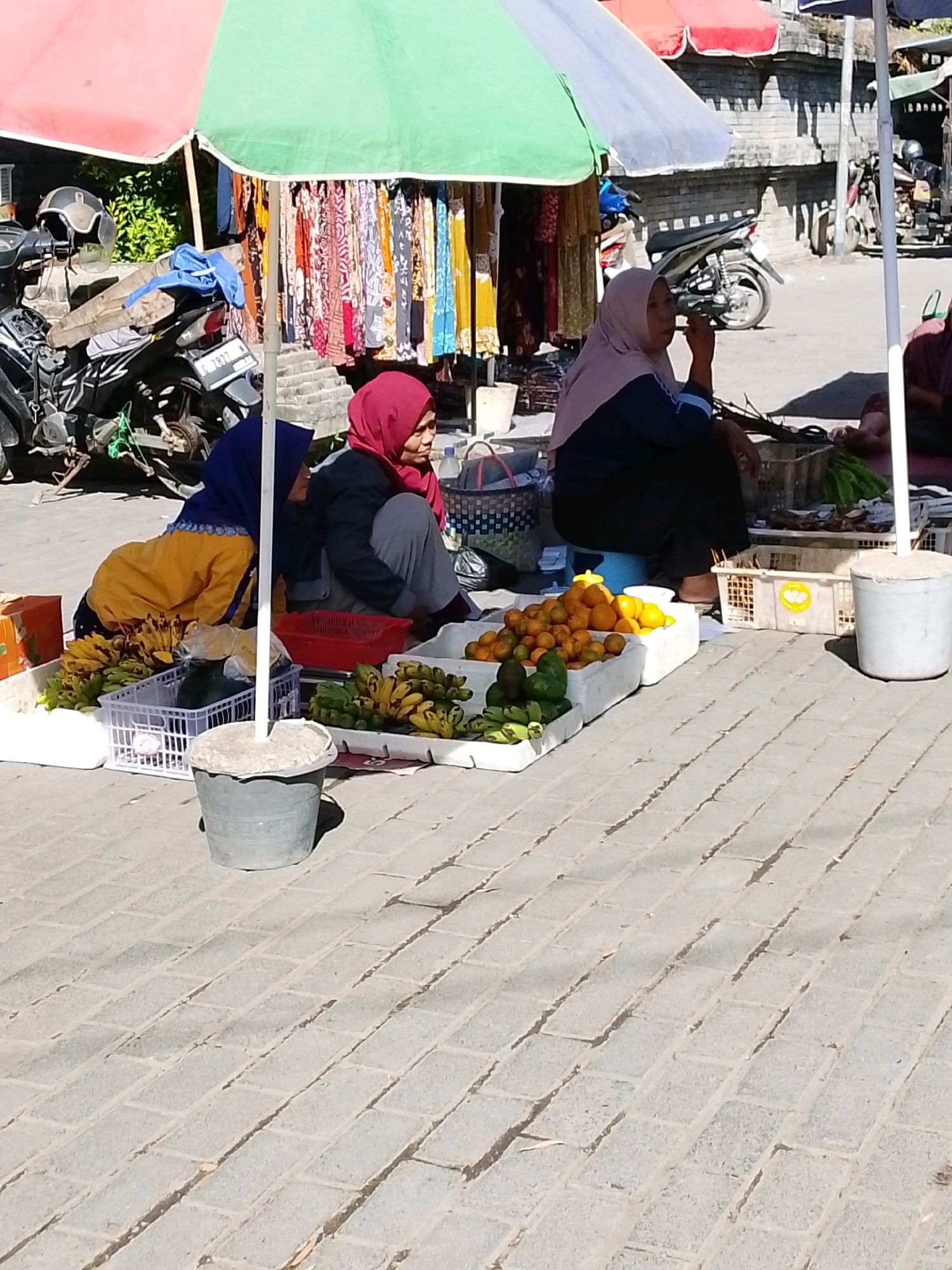 In Tegalwangi, Indonesia, under a red and green striped umbrella, three women sit on a paved walkway next to a street vendor stall. The woman on the left, wearing a blue hijab and a yellow top, is bent over and arranging fruit. The woman in the center, in a red hijab, is sitting and looking towards the right. The woman on the right, wearing a light pink hijab, is also seated and appears to be eating. In front of them are baskets and crates filled with bananas, oranges, and avocados. Behind them, clothing hangs from a rack, and a motorcycle is parked. A gray bucket rests on the ground in the foreground. The scene is illuminated by sunlight, casting shadows on the brick-paved ground.
