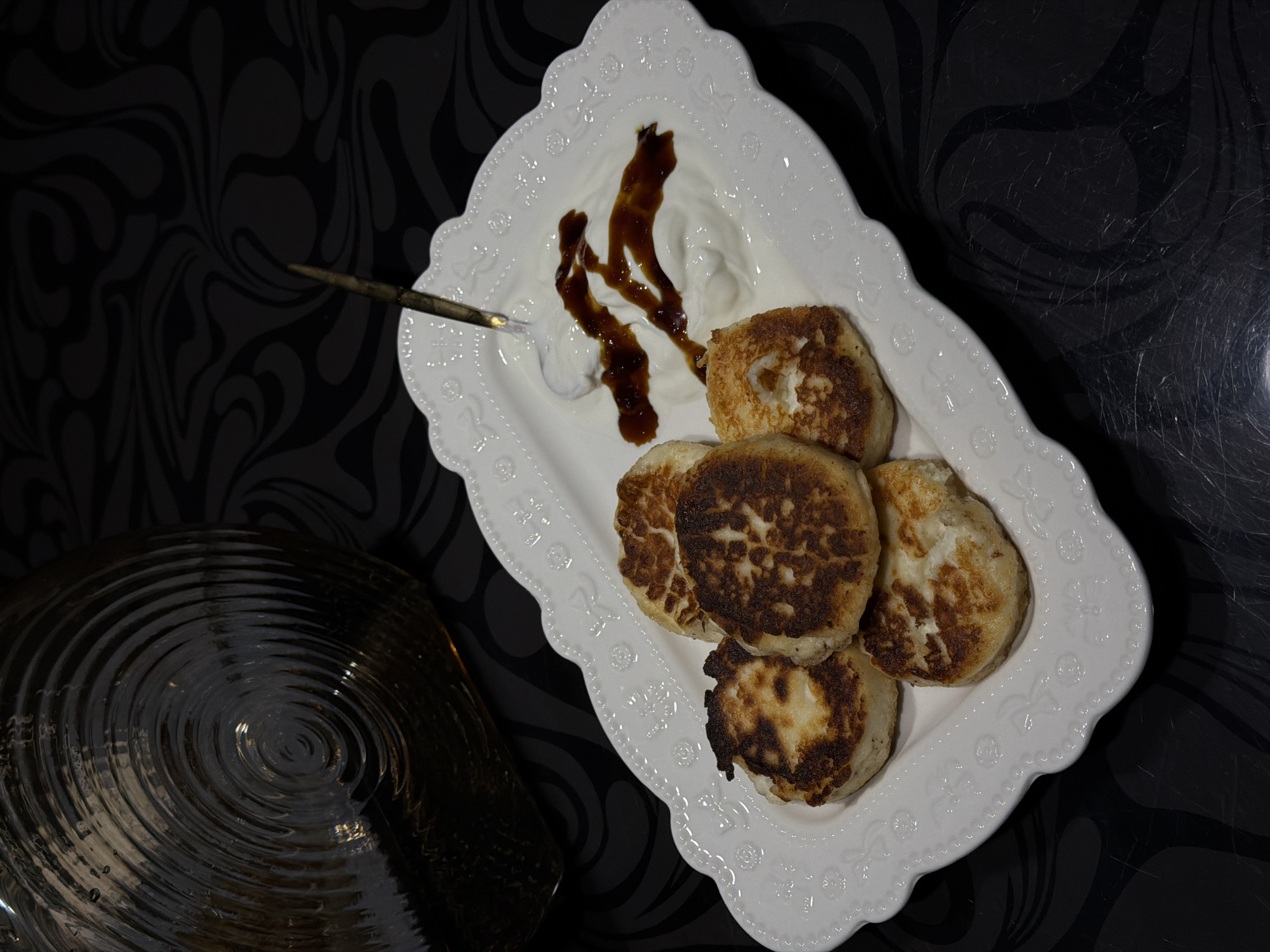 A high-angle shot depicts a plate of fried syrniki (cheese pancakes) with sour cream and syrup on a patterned black surface. The plate is white with a decorative edge. Six syrniki are stacked on the bottom half of the plate. A dollop of sour cream topped with a streak of syrup is on the top of the plate. A spoon is partially visible, embedded in the sour cream. To the left of the plate is a dark, round object with concentric circles, possibly a plate or bowl. The black surface has a swirling pattern. The location is Rossiyskiy, Russia.