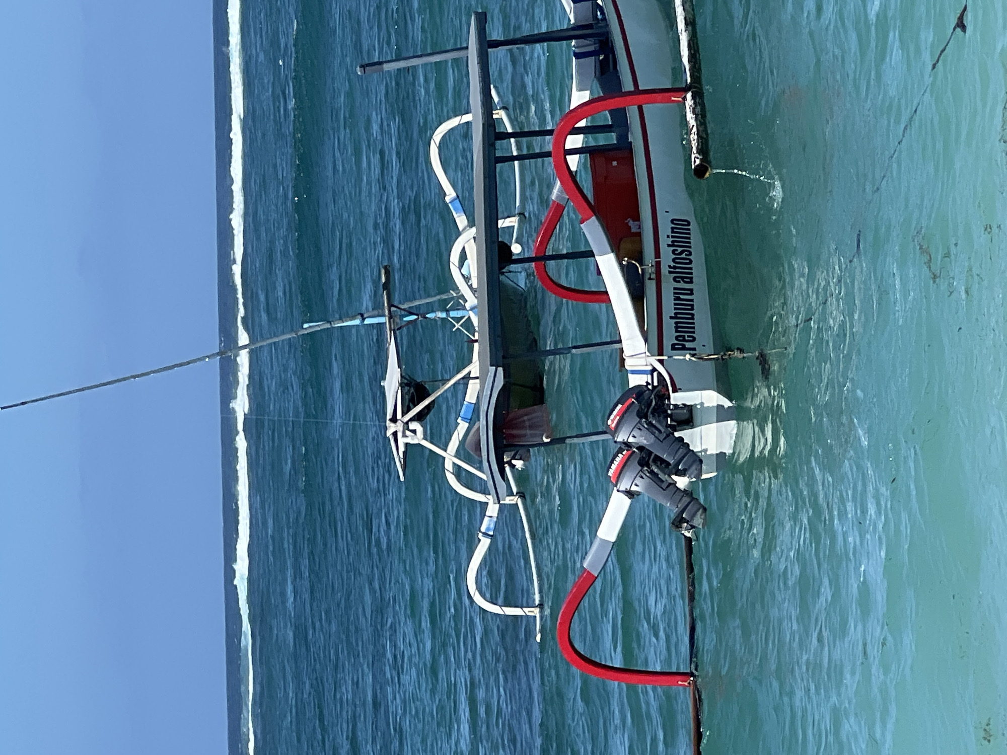 A white and red fishing boat with two Yamaha outboard motors is floating on turquoise water near the shore. The boat has a canopy for shade and outrigger stabilizers on both sides. The words "Pemburu alfoshino" are written on the side of the boat in black letters. In the background, waves are breaking on a reef. The sky is clear and blue. This scene is typical of the coastal waters in Kuta Selatan, Bali, Indonesia, where such traditional fishing boats are commonly used.