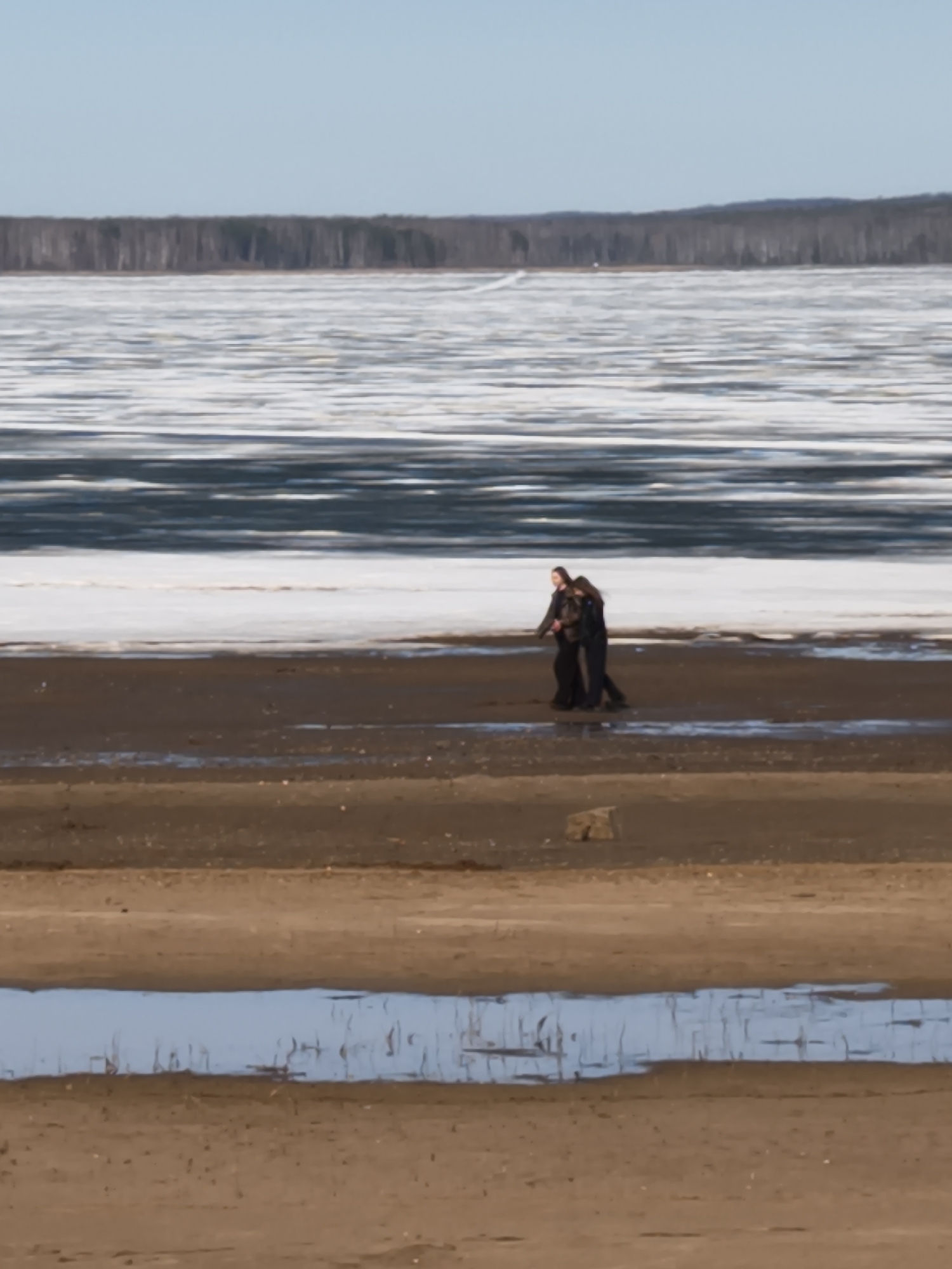 The photograph depicts a landscape with two people walking along a shoreline. The foreground features a sandy beach with a shallow pool of water reflecting the sky. The middle ground shows a partially frozen body of water, with patches of ice and open water. Two individuals are walking along the sandy shoreline near the water's edge. In the background, a treeline is visible against a clear sky. The location appears to be a natural setting, possibly near a lake or river in an unidentified city. The actions observed are individuals walking in a natural environment.
