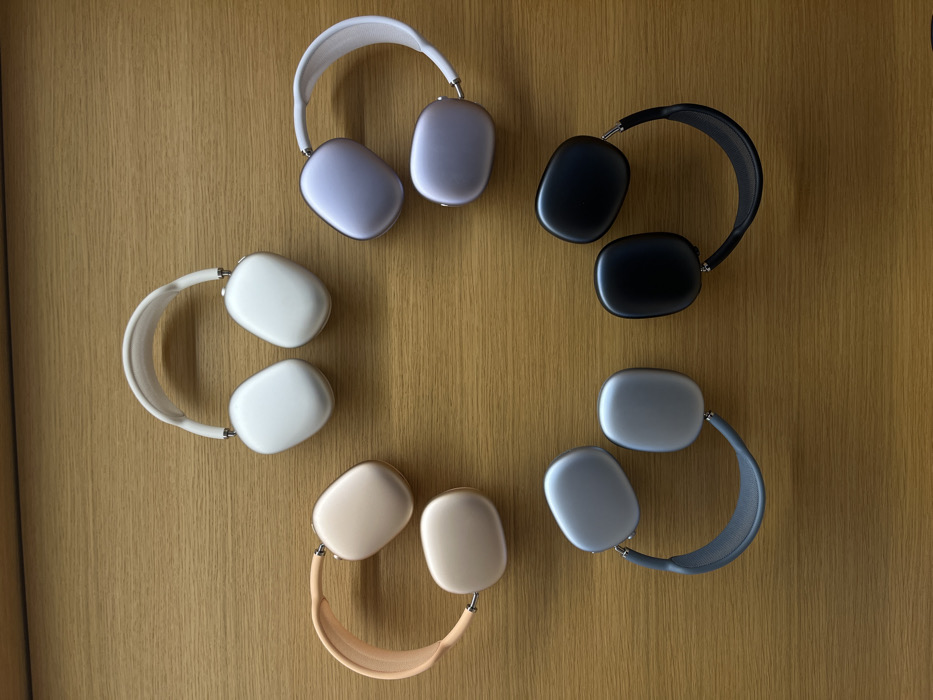 The image displays five pairs of Apple AirPods Max headphones arranged in a semi-circular pattern on a light-brown wooden surface. The headphones are presented in their various color options: from top-left clockwise, they appear to be Pink (Rose Gold), Silver, Purple (Sky Blue), Space Black, and Space Gray. Each pair is laid flat with the earcups facing upwards and the headbands slightly curved. The wooden surface is smooth and clean, consistent with a retail display table. No individuals are present in the scene. The overall environment suggests an indoor retail setting, specifically an Apple Store, given the proprietary products and display style. While the exact Apple Store in Paris, France, cannot be definitively identified from this image alone, the scene is characteristic of such a location, like the Apple Store Opéra or Champs-Élysées.
