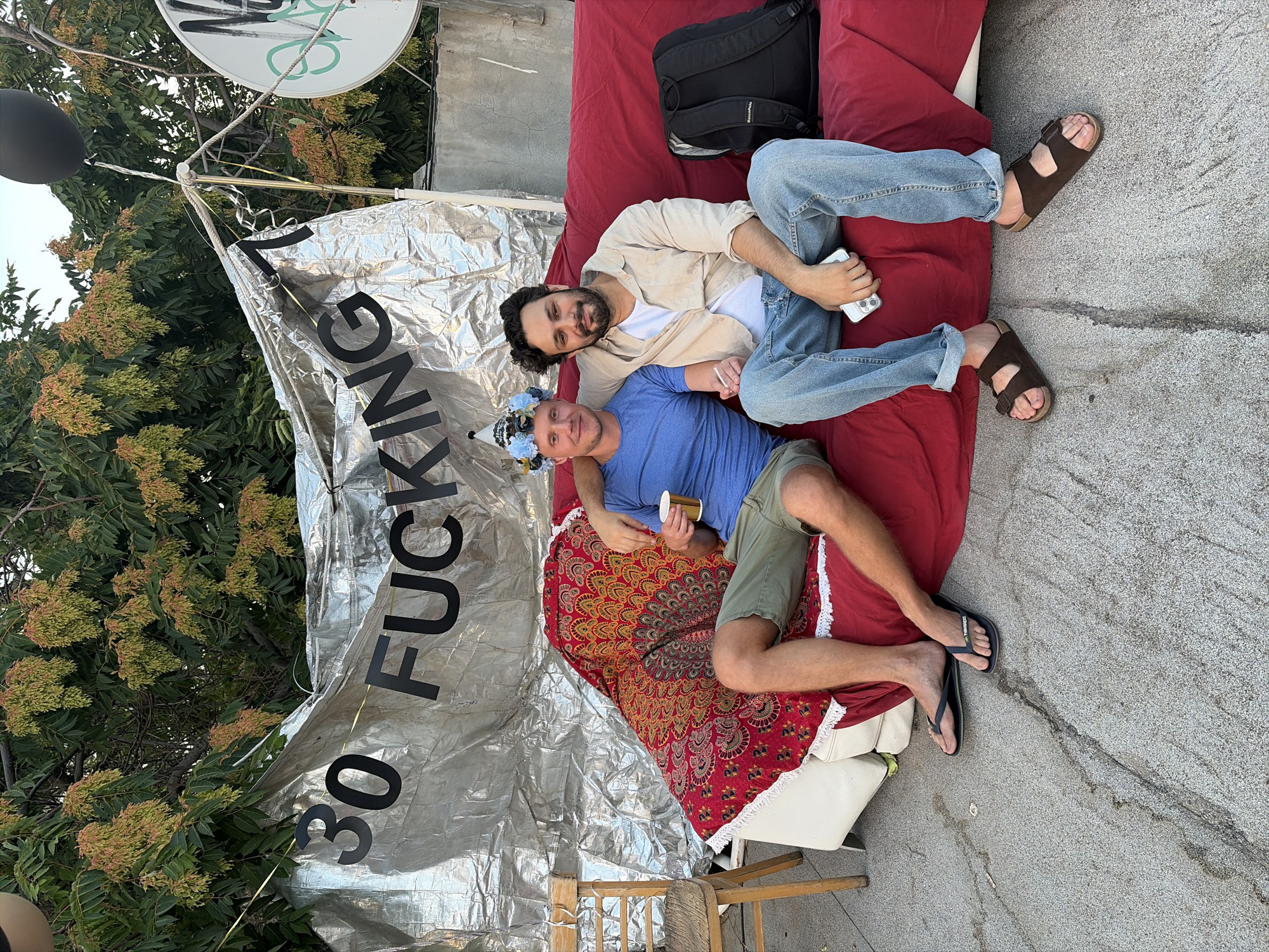 Two men are seated on a red couch outdoors. The man on the left wears a blue t-shirt, green shorts, and flip-flops. He has a paper cup in his right hand and a party hat on his head. The man on the right wears an open tan button-down shirt over a white t-shirt, blue jeans, and brown sandals. He holds a smartphone in his right hand. Behind them is a silver banner with black lettering that reads "30 FUCKING 7". A red blanket with a mandala pattern is draped over the couch. A black backpack rests on the right side of the couch. To the left of the couch is a wooden chair with a woven seat. The ground is paved with concrete that has cracks. Trees with seed pods are visible in the background. A black balloon is tied to a pole in the upper right corner, and a white circular object with graffiti is also visible. The scene appears to be in Tel-Aviv, Israel, likely a residential area with an outdoor space.