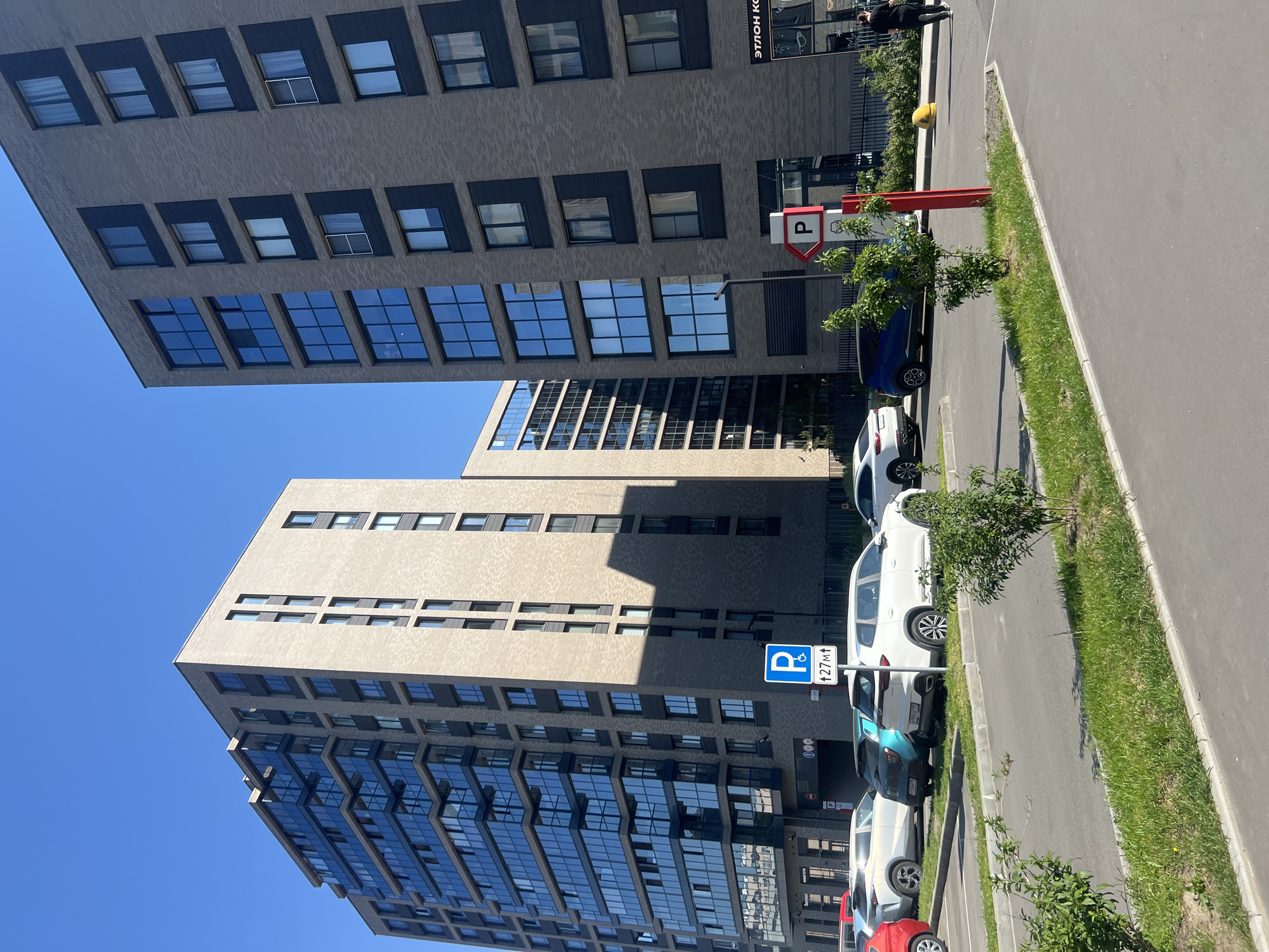 This image presents an urban scene dominated by two modern, multi-story buildings under a clear blue sky on a bright, sunny day. The building on the left is tall, featuring a light beige, textured facade with numerous dark-framed windows, many of which reflect the sky. Its upper sections recede, creating a layered appearance. To its right, a slightly shorter building with a dark grey brick-like exterior stands, also characterized by multiple dark-framed windows, some appearing open.
In the foreground and between the buildings, an asphalt parking lot is visible, with several cars parked. These include a white SUV, a blue sedan, a teal SUV, and a red car, among others. Green grass strips with small trees and bushes line the parking area, bordering a paved sidewalk or road.
Notable signs include a prominent red and white parking sign with a "P" symbol and a right-pointing arrow, positioned near the dark grey building. Further down, a blue and white parking sign with a "P" and a disabled access symbol shows the text "↑27м↓". On the facade of the dark grey building, the partial text "ЭТЛОН КО..." is visible, likely referring to "Etalon Comfort" or a similar development, fitting the Saint Petersburg context. A single person is faintly visible near the entrance of the dark grey building. The distinct shadows indicate it is daytime, likely mid-morning or afternoon.