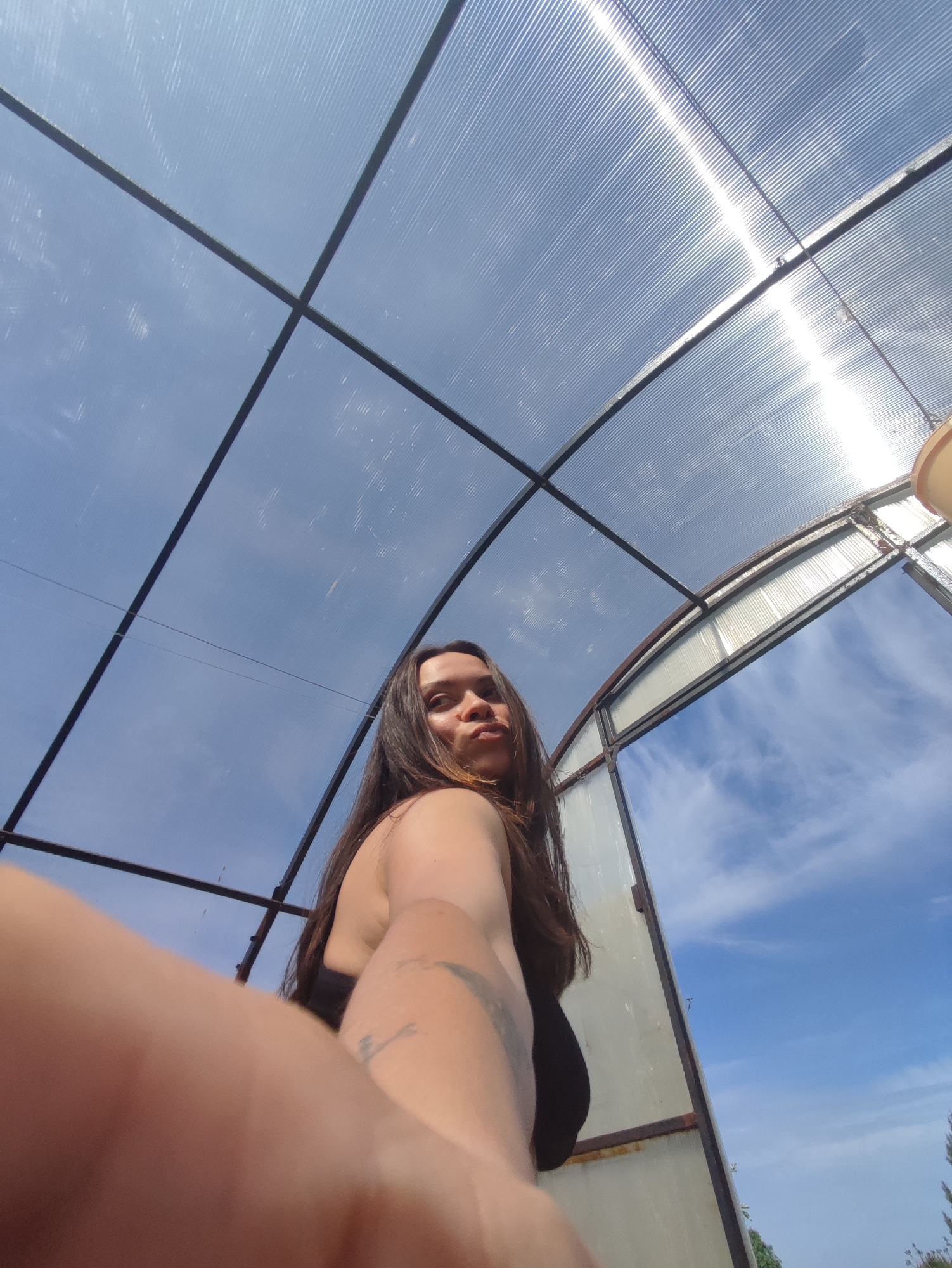 A low-angle, medium shot depicts a person with long dark hair inside a greenhouse, likely self-photographed using their extended arm. The person is wearing a dark top. The greenhouse structure is made of a metal frame covered with a transparent corrugated plastic material. The sky is visible through the structure, appearing partly cloudy. The location is unknown and cannot be determined from the visual information provided. The city name, Фоки, Russia, cannot be confirmed as the location.