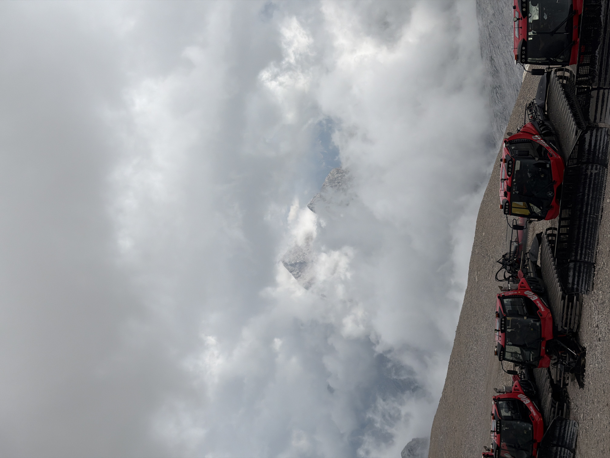 In Garmisch-Partenkirchen, Germany, on a rocky, mountainous terrain under a cloudy sky, four red snow grooming vehicles are parked in a line. The vehicles, with their tracks visible, are positioned on a gravelly surface. In the background, a mountain peak emerges from the thick clouds, its summit sharp and rocky. The overall scene depicts a rugged, high-altitude environment.