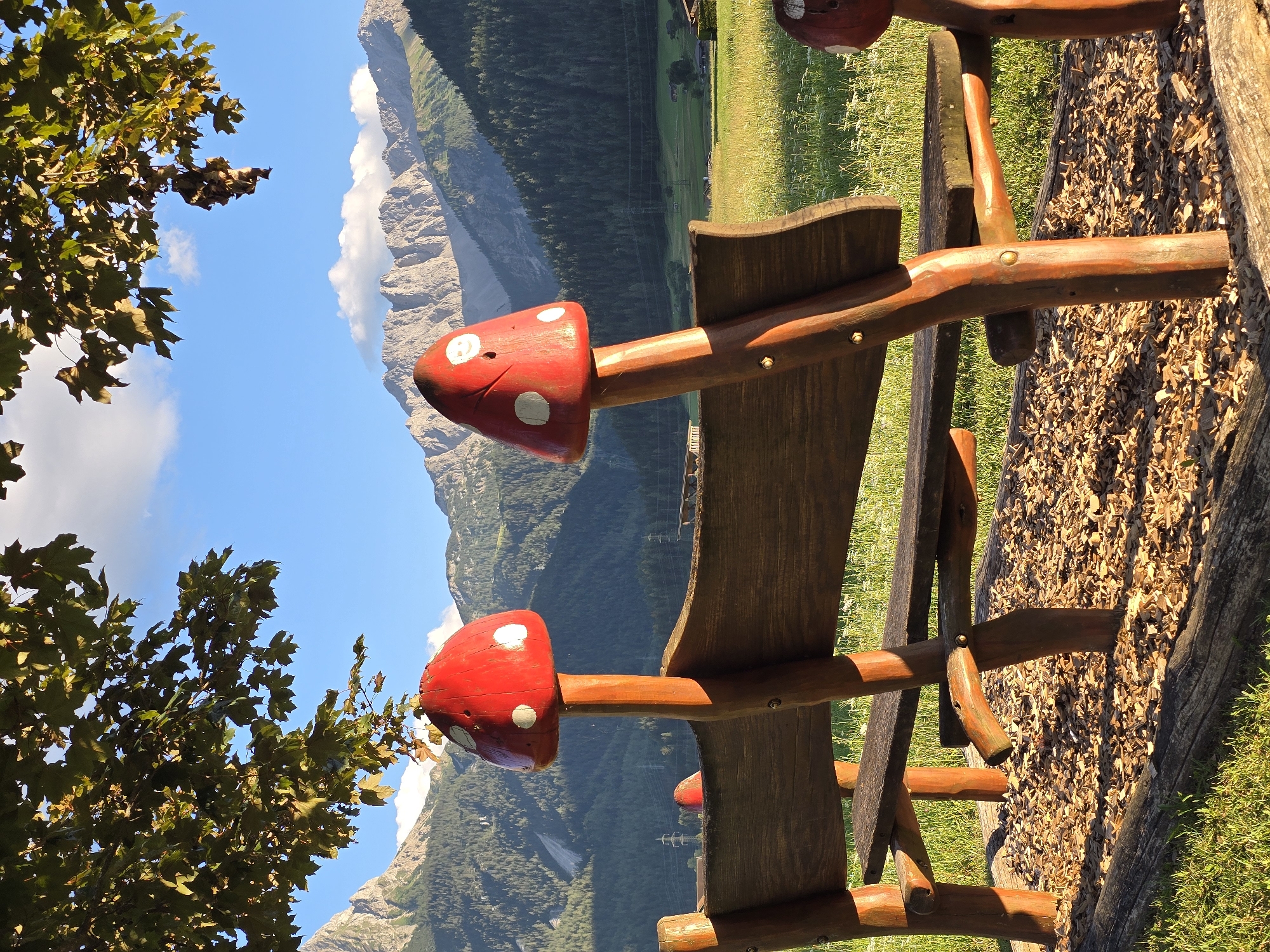 The image depicts a wooden park bench with two decorative mushroom-shaped supports, set against a backdrop of the Austrian Alps in Lermoos. The bench is constructed from rough-hewn wood, with two mushroom caps painted red with white spots serving as the backrest supports. A third, smaller mushroom-shaped support is visible to the right. The ground around the bench is covered with wood chips and green grass. In the background, a sun-drenched meadow stretches towards a dense forest that climbs the lower slopes of majestic, rugged mountains. The sky above is blue with scattered white clouds, and the branches of a large tree overhang the scene from the top left. The lighting suggests it is late afternoon, casting long shadows. There are no people visible in the image.