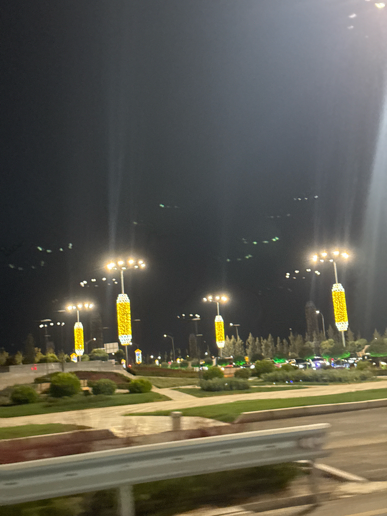 The image depicts a night scene in Tashkent, Uzbekistan, likely on a wide boulevard or park area. Tall, decorative lampposts with bright yellow lighting illuminate the darkness. These lampposts are prominent features, some with intricate, cylindrical yellow and white decorations. In the background, trees and foliage line the area, and distant car lights suggest ongoing traffic. The overall impression is one of a well-lit urban environment, possibly a newly developed or recently renovated public space, designed for aesthetic appeal at night. The ground appears to be paved and landscaped with grass and low bushes. The image is taken from a moving vehicle, as evidenced by the slight blur and the railing in the foreground.