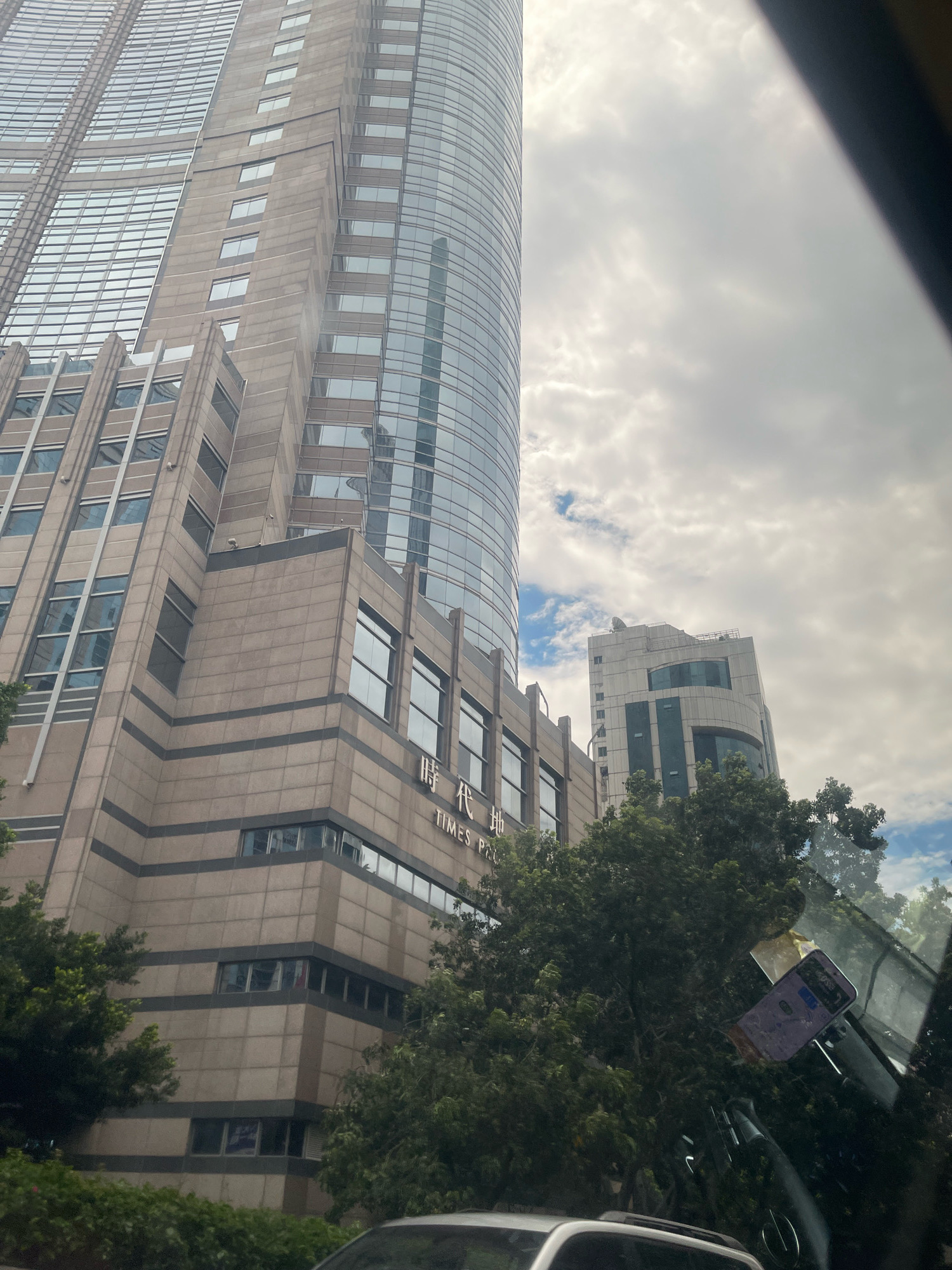 The image shows a low-angle, exterior view of tall, modern buildings against a cloudy sky. The prominent building in the foreground is clad in beige and brown panels with dark horizontal stripes and numerous rectangular windows. A portion of a taller, glass-fronted skyscraper is visible to the left, reflecting the sky. The building in the foreground has signage that reads "TIMES PA" in English and Chinese characters. In the background, another multi-story building with a lighter facade and dark-tinted windows is visible. Lush green trees and foliage are in the foreground and middle ground, obscuring the lower portions of the buildings. A car's interior is partially visible in the lower right corner, with a purple electronic device attached to the windshield. The scene appears to be in an urban environment, possibly the Yuexiu District of China, based on the provided context. No people are directly visible in the image.