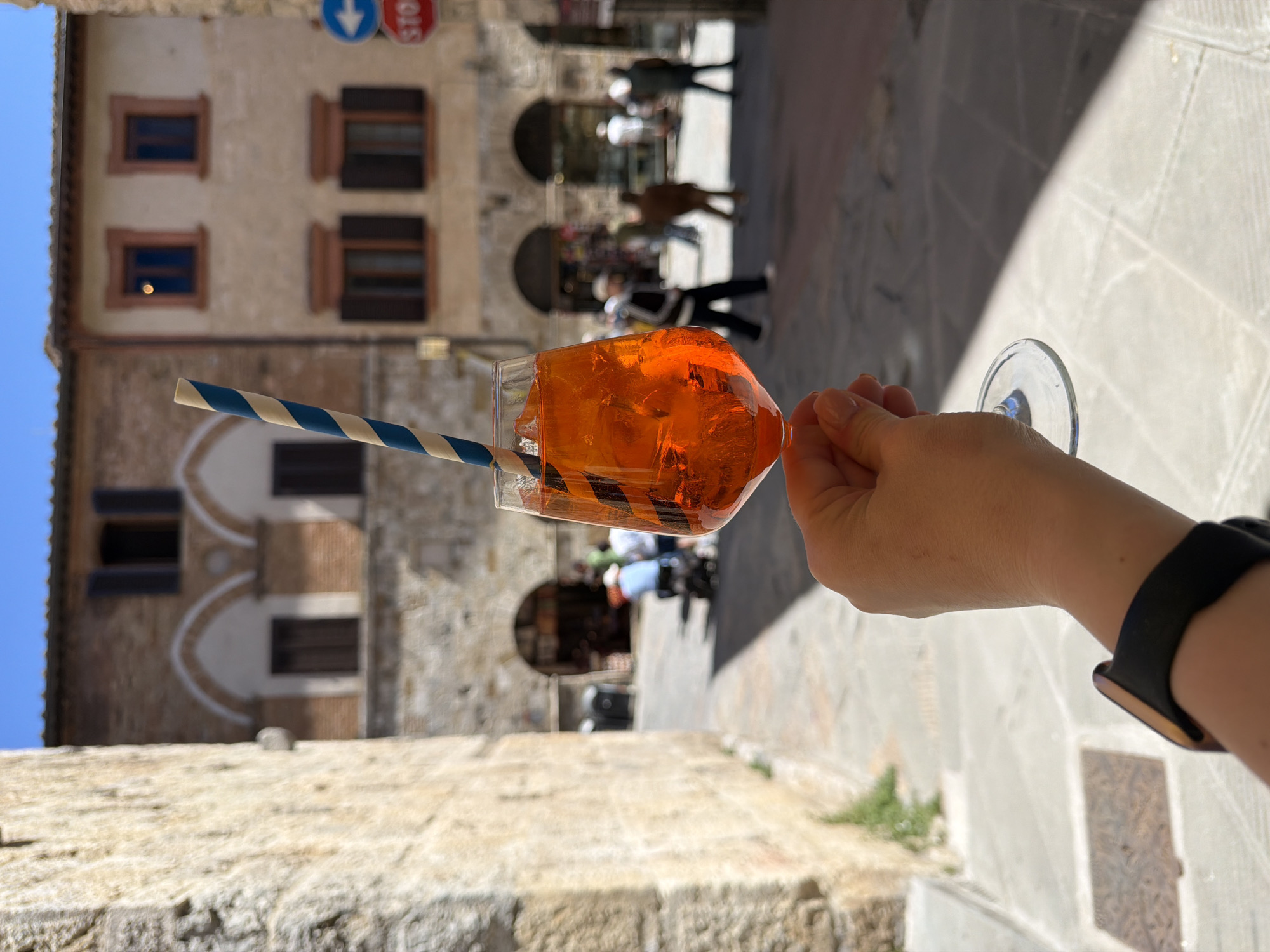 The image, when viewed upright, presents a first-person perspective of a hand holding a refreshing orange-colored drink. The drink, likely an Aperol Spritz, is served in a stemless wine glass filled with ice cubes and features a blue and white striped paper straw. The hand, with manicured fingernails, has a black smartwatch on its wrist.
The scene unfolds on a sunny day in what appears to be a historic European town, specifically San Gimignano, Italy, as indicated by the location context. In the background, traditional stone buildings with multiple windows line a narrow street. The windows feature dark brown wooden shutters, some open and some closed. Arched doorways or passages are also visible within the building facades.
Blurred figures of people can be seen walking further down the street, adding to the bustling atmosphere of the town. Strong sunlight casts distinct shadows on the stone ground, highlighting the clear and bright weather conditions. Visible details include a blue circular traffic sign with a white downward arrow and a partially visible red octagonal "STOP" sign mounted on one of the buildings. A clear blue sky is visible at the top left of the frame. The image conveys a sense of relaxation and enjoyment in a picturesque, historical setting.