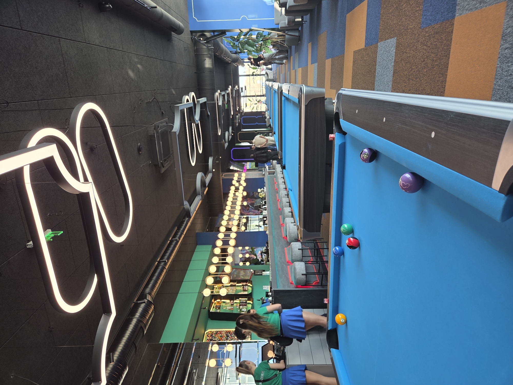 The image depicts an interior space featuring a row of pool tables. Two young women in green shirts and blue skirts are playing pool. The walls are decorated with illuminated graphic elements and there are additional gaming areas further down the space. The scene appears to be in a modern entertainment venue, possibly in the city of Varna, Bulgaria.