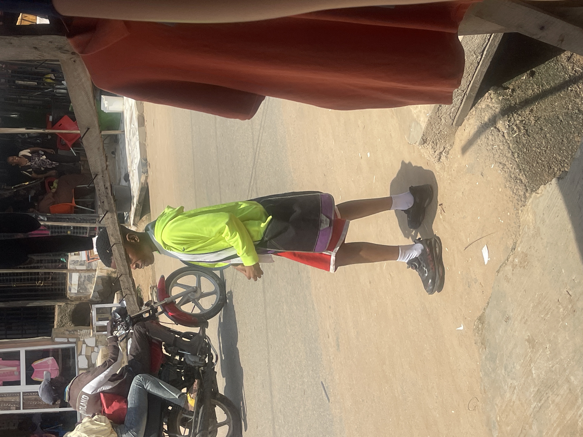 In the city of Abuja, Nigeria, a young Black male walks on a dusty, unpaved street. He wears a neon green hooded jacket over a white t-shirt, red shorts, black athletic shoes with white socks, and a black crossbody bag with purple and white accents. His hair is short and neat. To his left, a motorcyclist wearing a dark brown jacket with "ENGLAND" written in white across the back, a blue t-shirt, a gray baseball cap, and dark pants drives a red motorcycle. The motorcyclist is carrying a passenger, a Black female wearing a light yellow top and blue jeans, who is seated behind him. In the background, a market scene unfolds with stalls and people. An orange garment hangs on a wooden structure in the foreground, partially obscuring the view of the street. The overall environment suggests a bustling urban setting with elements of both commerce and everyday life.