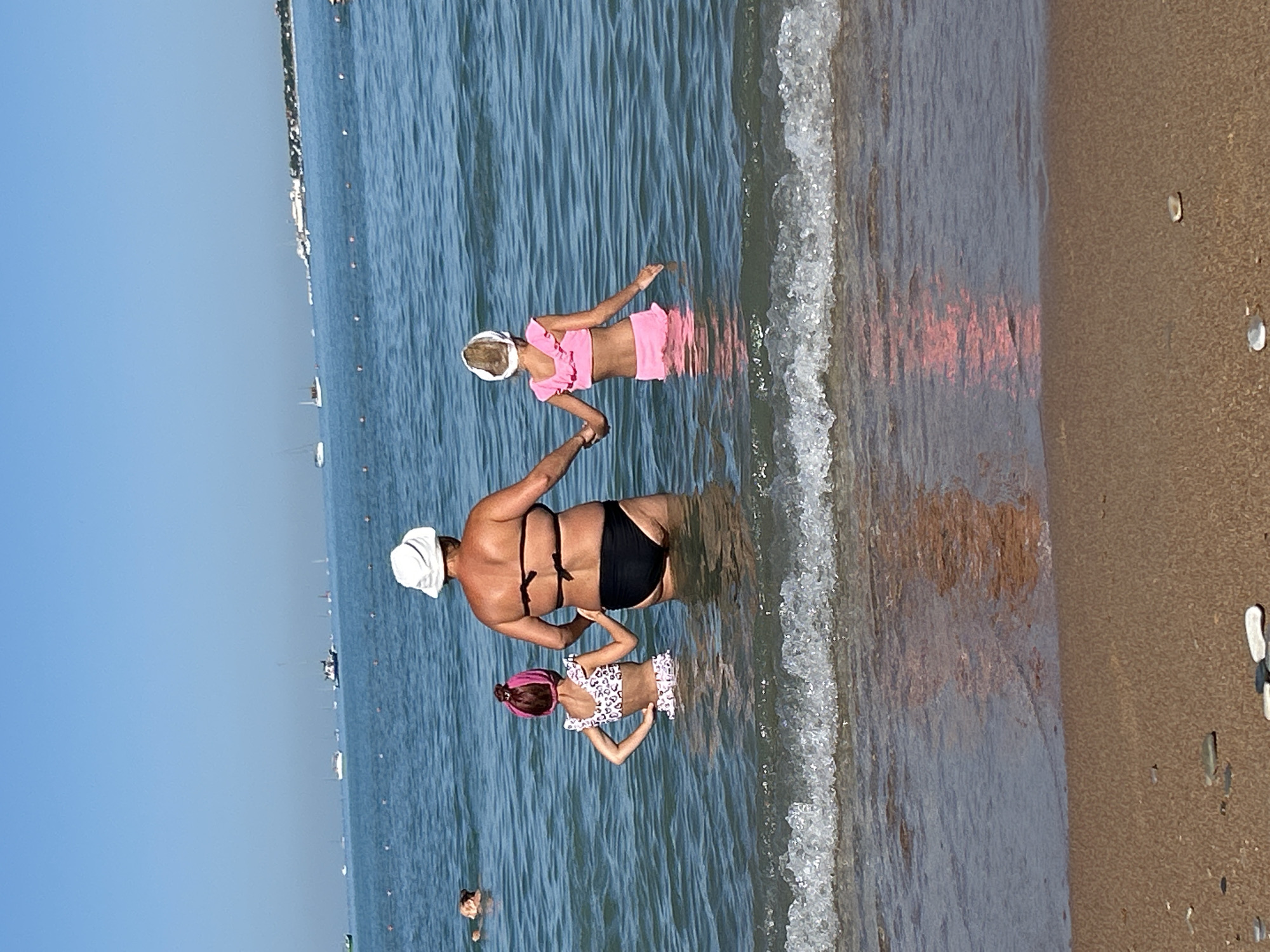 In the Black Sea off the coast of Gelendzhik, Russia, a woman wearing a white sun hat and black bikini stands waist-deep in the water. She is holding hands with two children. The child on her left wears a pink and white patterned bikini and a pink hairband. The child on her right wears a pink two-piece swimsuit and a white head wrap. In the background, several boats are visible on the water, and a distant shoreline with buildings and trees can be seen. The foreground shows the sandy beach with a gentle wave breaking.