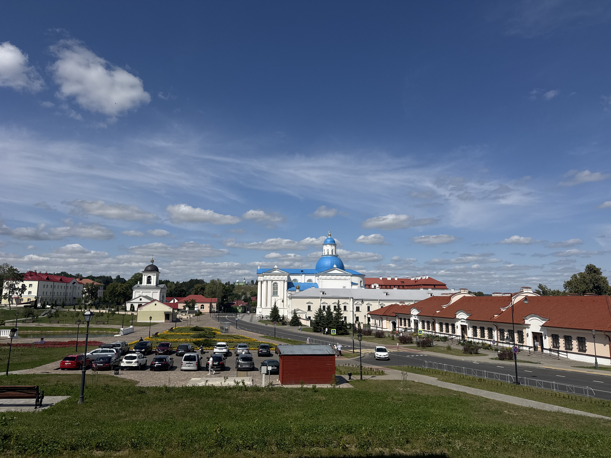 The image shows a wide view of a town square or plaza in Zhyrvicky Rural Council, Belarus. In the foreground, there is a grassy slope with a park bench. Beyond the slope, a paved area has a parking lot with approximately 15 cars. Adjacent to the parking lot is a small red shed and a flower bed with yellow and red flowers. Across the paved area, there is a road with several cars.
Dominating the midground and background are several buildings. To the left, a white building with a dark steeple and a bell tower stands prominently. Further to the right is a large white building with a bright blue dome, likely a church or cathedral. Next to this is a long, low-rise building with a red tiled roof, featuring multiple windows and arched doorways. More buildings with red roofs are visible further in the background, interspersed with trees. The sky above is a bright blue with scattered white clouds. The overall scene suggests a sunny day in a European town with historical architecture.