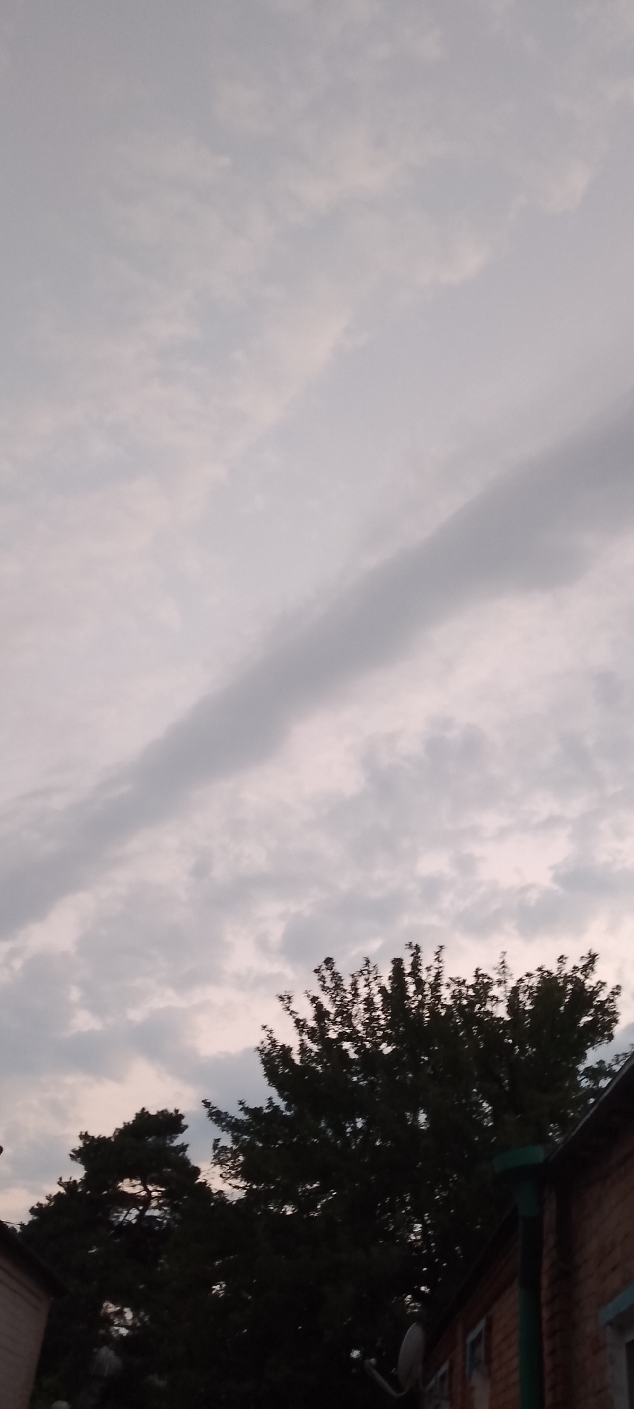 The image depicts a cloudy sky with a large, dark cloud band stretching diagonally across the frame. In the lower portion of the image, silhouetted against the lighter sky, are the dark shapes of trees and the roofline of a brick building. The scene appears to be an outdoor view from a residential area, possibly a backyard or alleyway, in Belgorod, Russia, given the architectural style of the building and the type of vegetation. No people are visible. The lighting suggests it is either early morning or late evening.