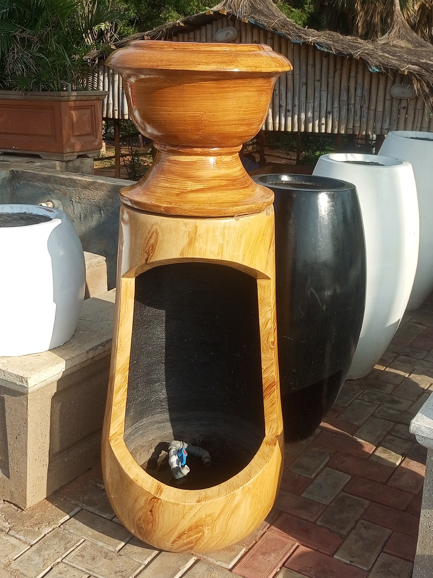 The image shows an outdoor scene, likely a market or garden center, in Gombe, Nigeria. The primary subjects are several large planters. In the foreground, a tall, uniquely shaped planter is made of what appears to be wood or a wood-like material with a glossy finish. It has an opening at the bottom revealing a dark interior with plumbing fixtures, suggesting it might be a fountain or water feature.
Behind this planter, there are other large planters, including a glossy black one and two tall white ones. To the left, a rectangular brown planter is visible. In the background, a rustic structure with a thatched roof and bamboo walls provides a backdrop, hinting at a traditional or artisanal setting. Lush green foliage, possibly palm trees, can be seen behind the structure.
The ground is paved with interlocking bricks in shades of red and grey. The lighting suggests it is daytime, with strong shadows indicating a sunny day, possibly late morning or early afternoon. There is no visible text in the image. The overall impression is of a display of garden ornaments or pottery in a warm, possibly humid, climate.