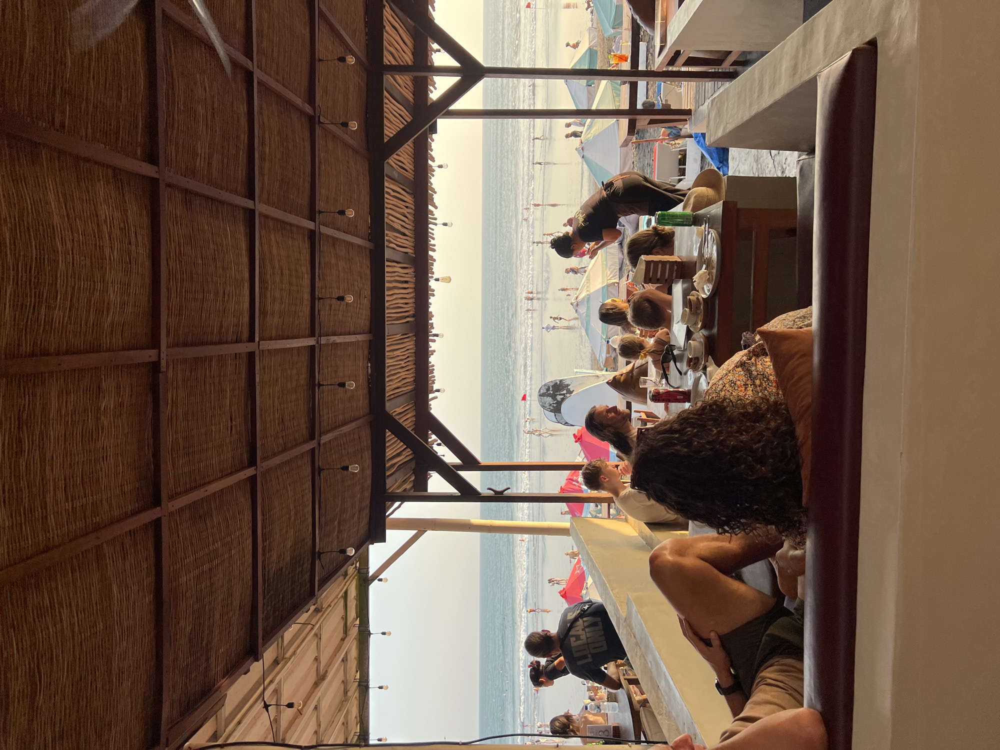 The image depicts a beachside restaurant in North Kuta, Indonesia, at sunset. A thatched roof covers an open-air dining area where several people are seated at tables. In the foreground, patrons are seen from behind, relaxing on a couch. In the midground, a server in a black uniform attends to a table. Beyond the restaurant, a crowded beach is visible with people walking and swimming in the ocean. Umbrellas dot the sand, and surfboards are propped up against a railing. The warm, golden light of the setting sun casts long shadows, creating a relaxed and vibrant atmosphere.