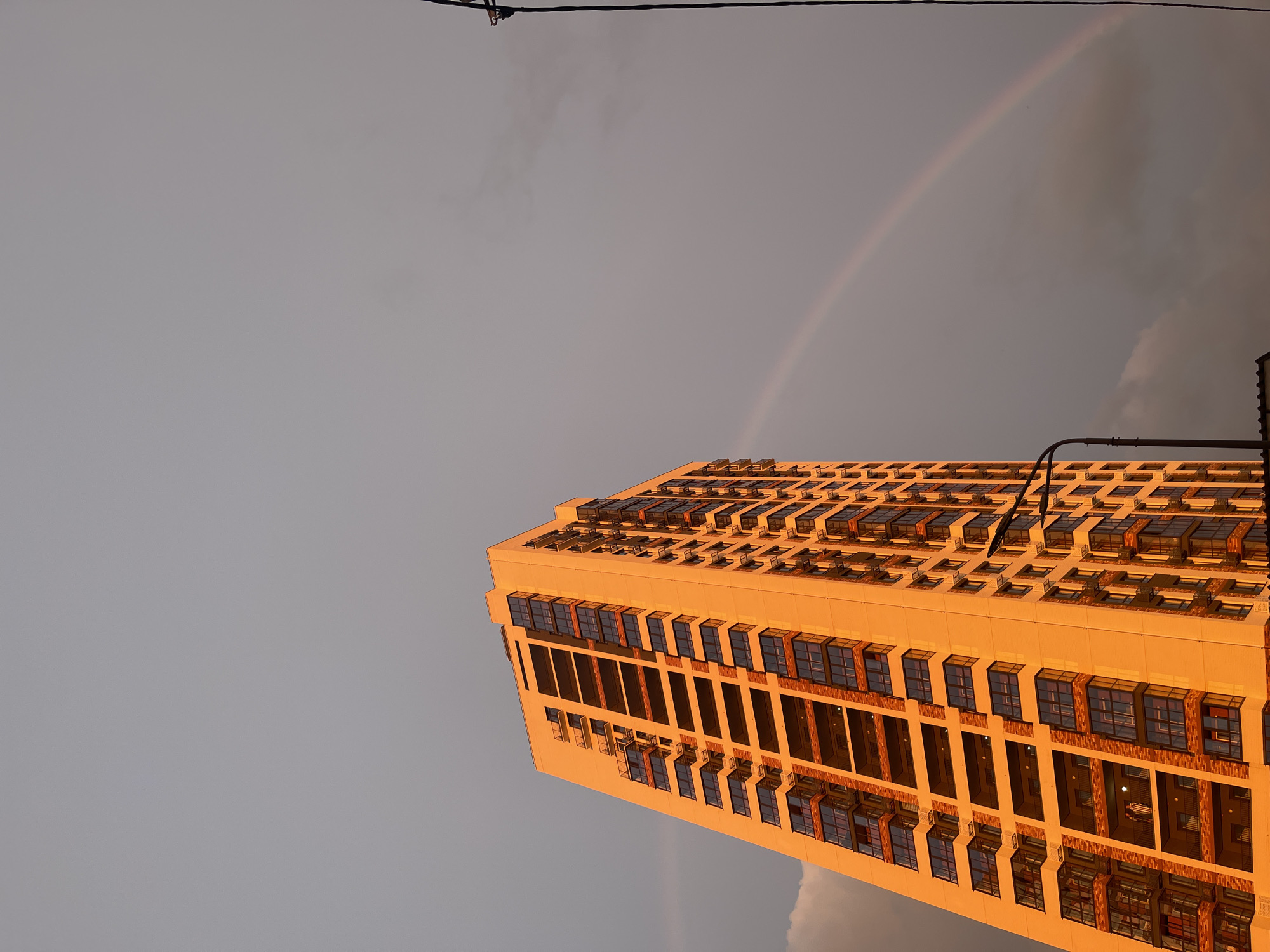The image displays a tall, modern building with a repetitive facade of windows, set against a cloudy sky. A faint rainbow arcs across the upper portion of the sky, suggesting recent rainfall or ongoing atmospheric conditions conducive to rainbow formation. The building's exterior is bathed in a warm, golden light, indicating that it is either sunrise or sunset. The sky itself is a muted gray, with some darker cloud formations visible towards the right side of the frame. A thin, dark power line runs horizontally across the very top of the image, with a small insulator visible. The building's architecture is characterized by numerous rectangular windows, some appearing to be open, and a pattern of vertical and horizontal divisions. The overall impression is one of an urban environment during a transitional period of the day, with a natural atmospheric phenomenon adding a touch of color. No people are visible, and no text is present in the image.