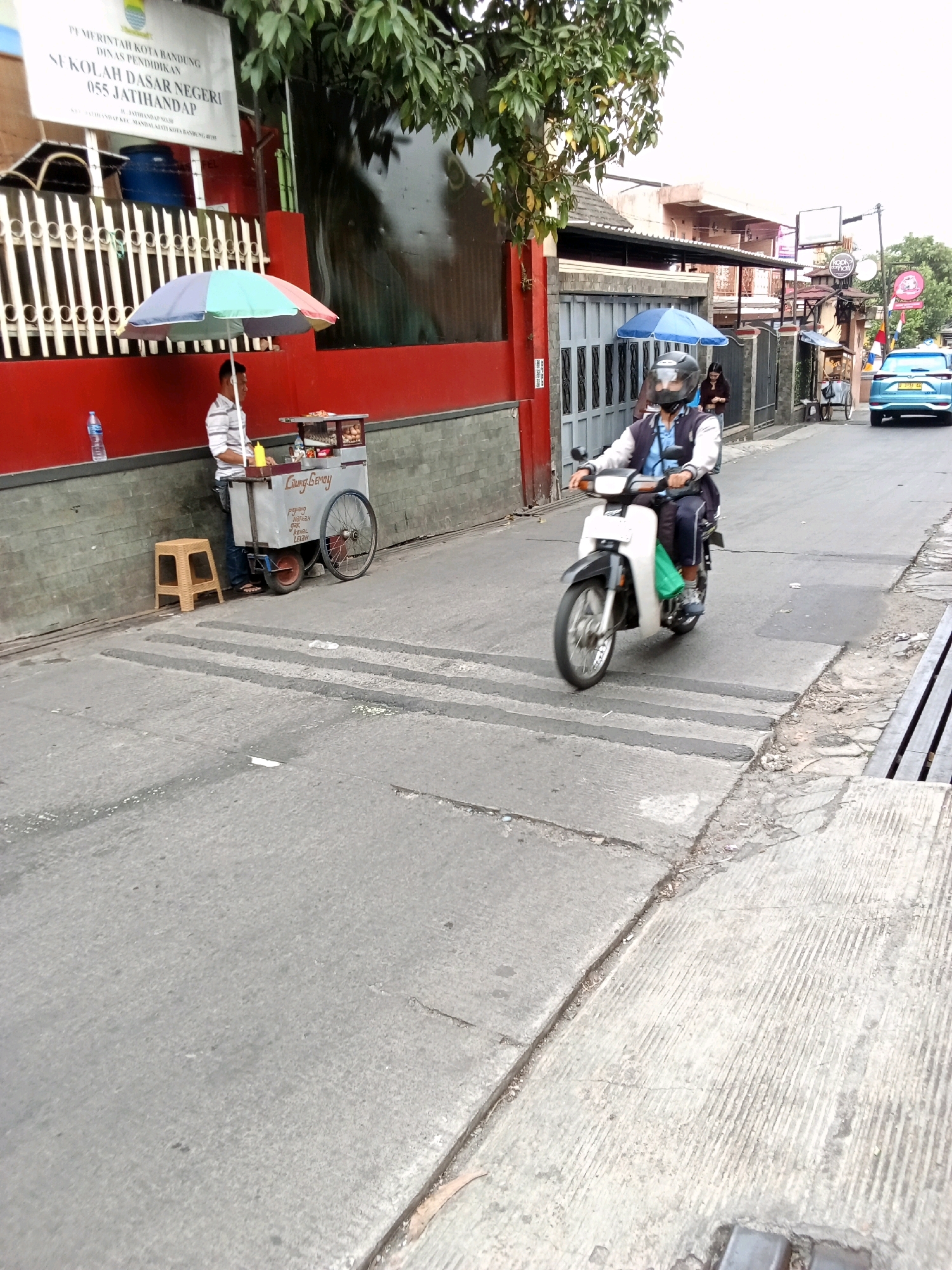 The image shows a street scene in Bandung City, Indonesia. A man is standing next to a food cart under a colorful umbrella, likely selling food. The cart has wheels and appears to be made of metal. A stool is placed next to the cart. In the background, a person is riding a motorcycle, wearing a helmet and a jacket. The street has a raised concrete pedestrian crossing. Buildings and trees line the street, and a blue car is visible further down the road. A sign with text in Indonesian is also visible on a building. The overall scene depicts a typical urban environment with commercial activity.