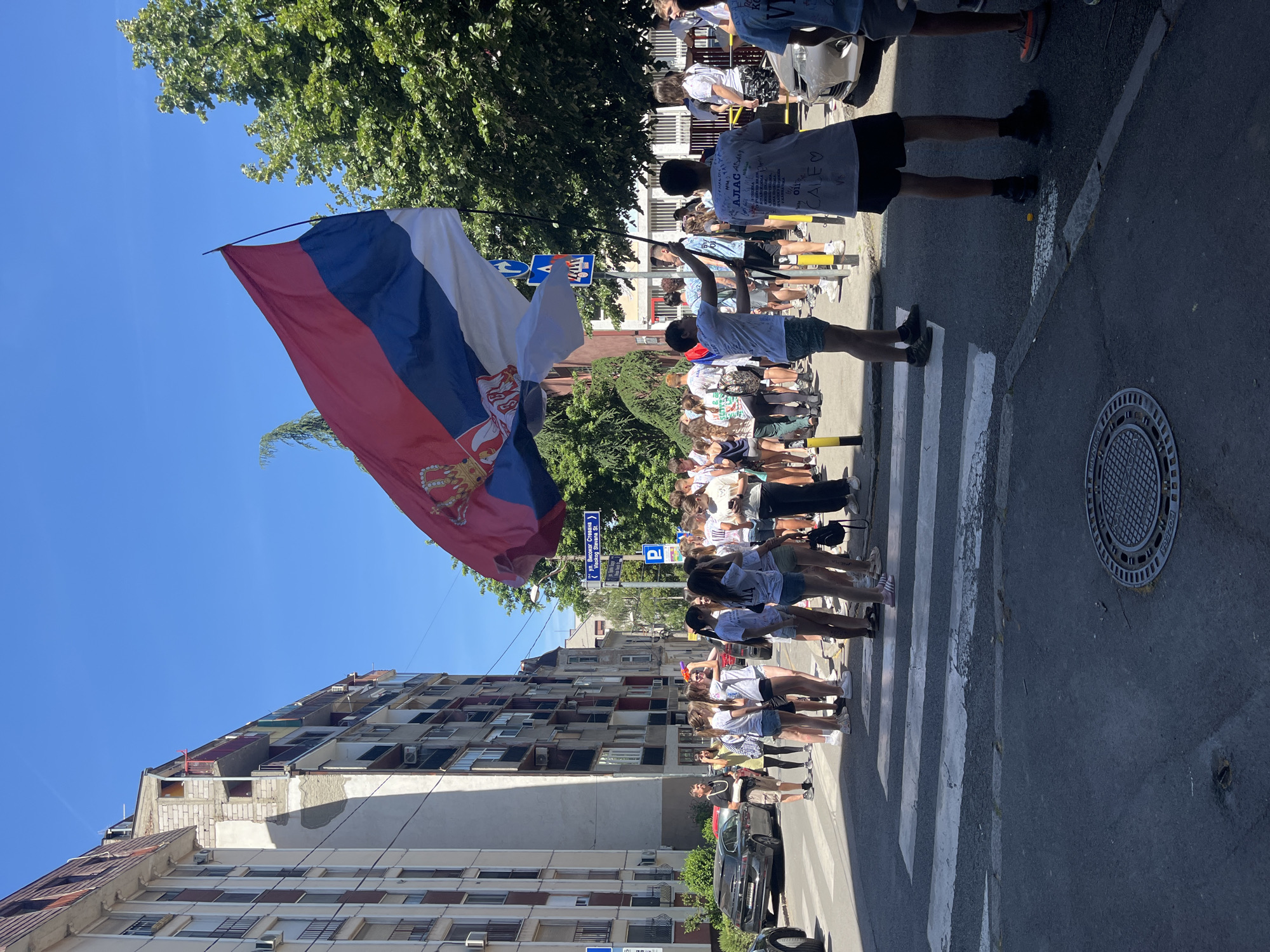 A street scene in Belgrade, Serbia, depicts a group of young people, many wearing white t-shirts with writing on them, gathered on a zebra crossing in front of a multi-story building. A large Serbian flag is prominently displayed. Cars are parked along the side of the road. Street signs are visible, including one indicating "Високог Стевана" (Visokog Stevana) and "Visokog Stevana St.". The sky is clear and blue. The scene appears to be a celebration or gathering of some kind.