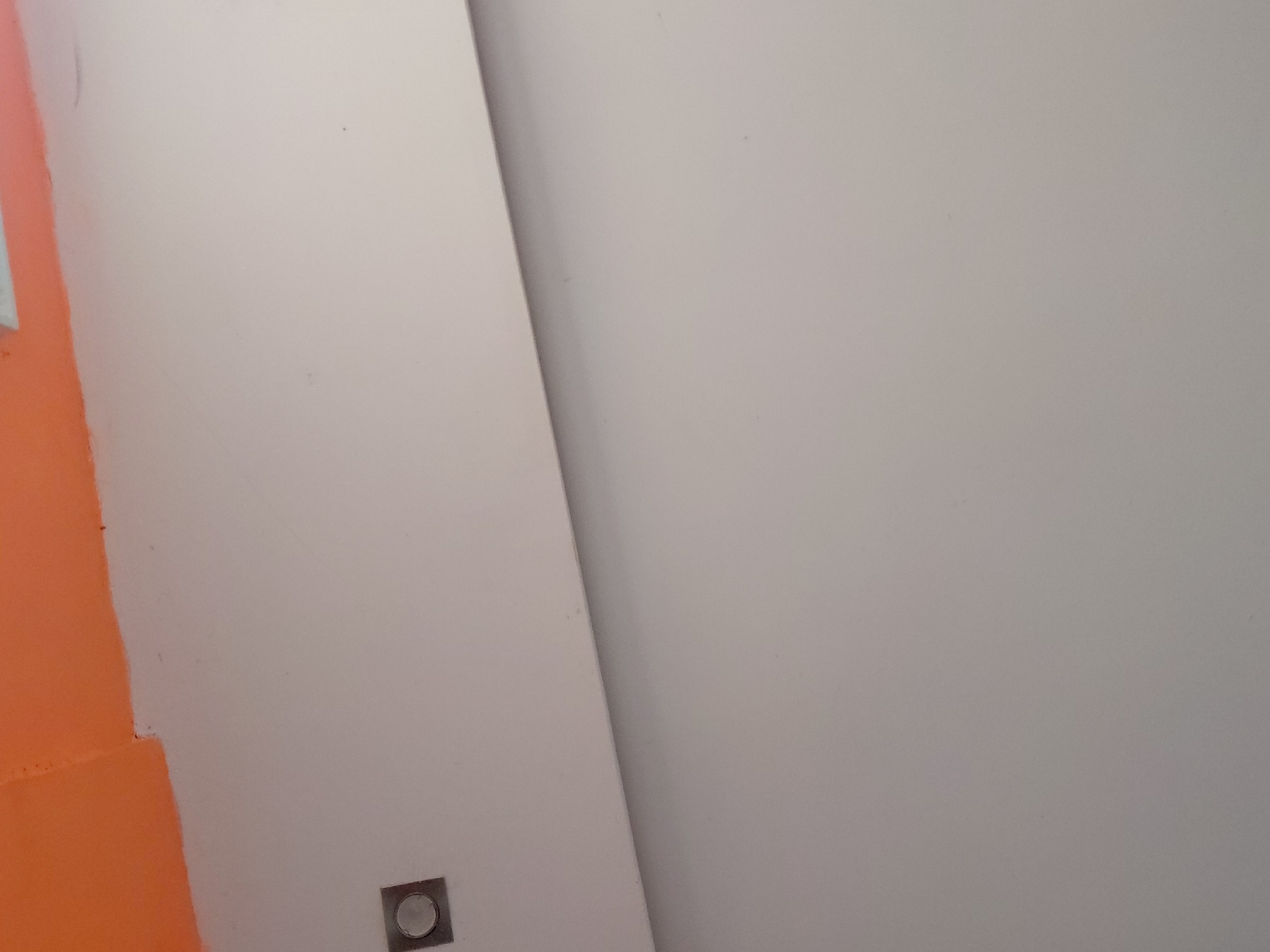The image displays an abstract close-up of an interior wall or surface, likely in Santa Ana, Venezuela. The scene is bisected by a prominent diagonal line, dividing a large light off-white or very light grey area into two planes, possibly indicating an interior corner or a seam between panels. To the far left, a vibrant orange section runs vertically, meeting the off-white surface with a slightly uneven, hand-painted edge.
Near the bottom-left corner of the off-white area, adjacent to the orange paint, there is a small, square object with a circular center. This object appears to be a recessed electrical outlet, button, or indicator, framed in metallic or light-colored plastic. The surfaces are smooth, suggesting painted plaster or drywall.
The lighting is even and diffused, casting no harsh shadows, which makes the time of day indeterminate. There are no people or specific activities depicted. No visible text is present. The composition emphasizes color contrast, geometric lines, and surface textures, possibly capturing a detail within a building interior during or after renovation.