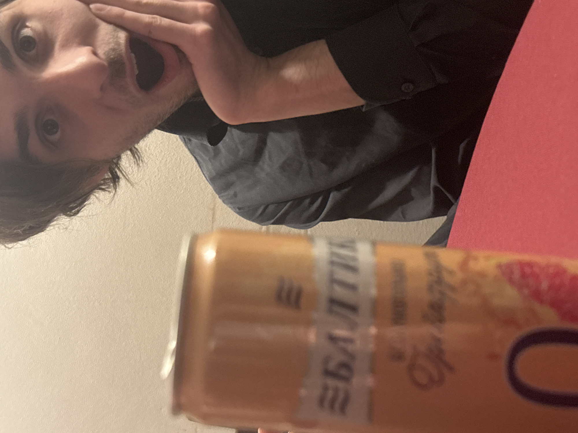 A close-up shot features a man with dark hair and light skin wearing a dark button-down shirt. He is holding his hand to his face, expressing surprise with a wide-open mouth. In the foreground, a blurred can of Baltika beer is visible, with the Cyrillic lettering identifying it. A red surface is seen on the right side of the frame. The background appears to be a plain wall. There is no information that specifies the location as Moscow, Russia.