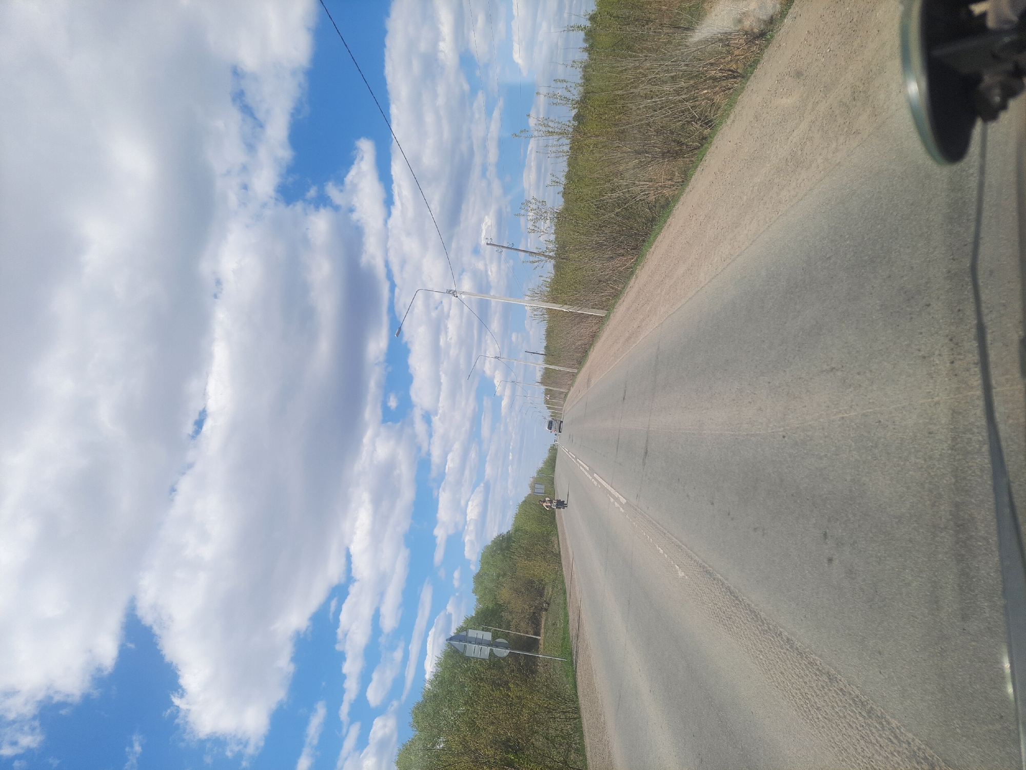 The image depicts a straight asphalt road stretching into the distance, likely taken from within a vehicle. A motorcyclist is visible in the distance, and a vehicle further ahead. Power poles line the left side of the road, with trees visible beyond them. The sky is blue with white, puffy clouds. The right side of the road has a more overgrown, grassy shoulder. The location is in Научный Городок, Russia.