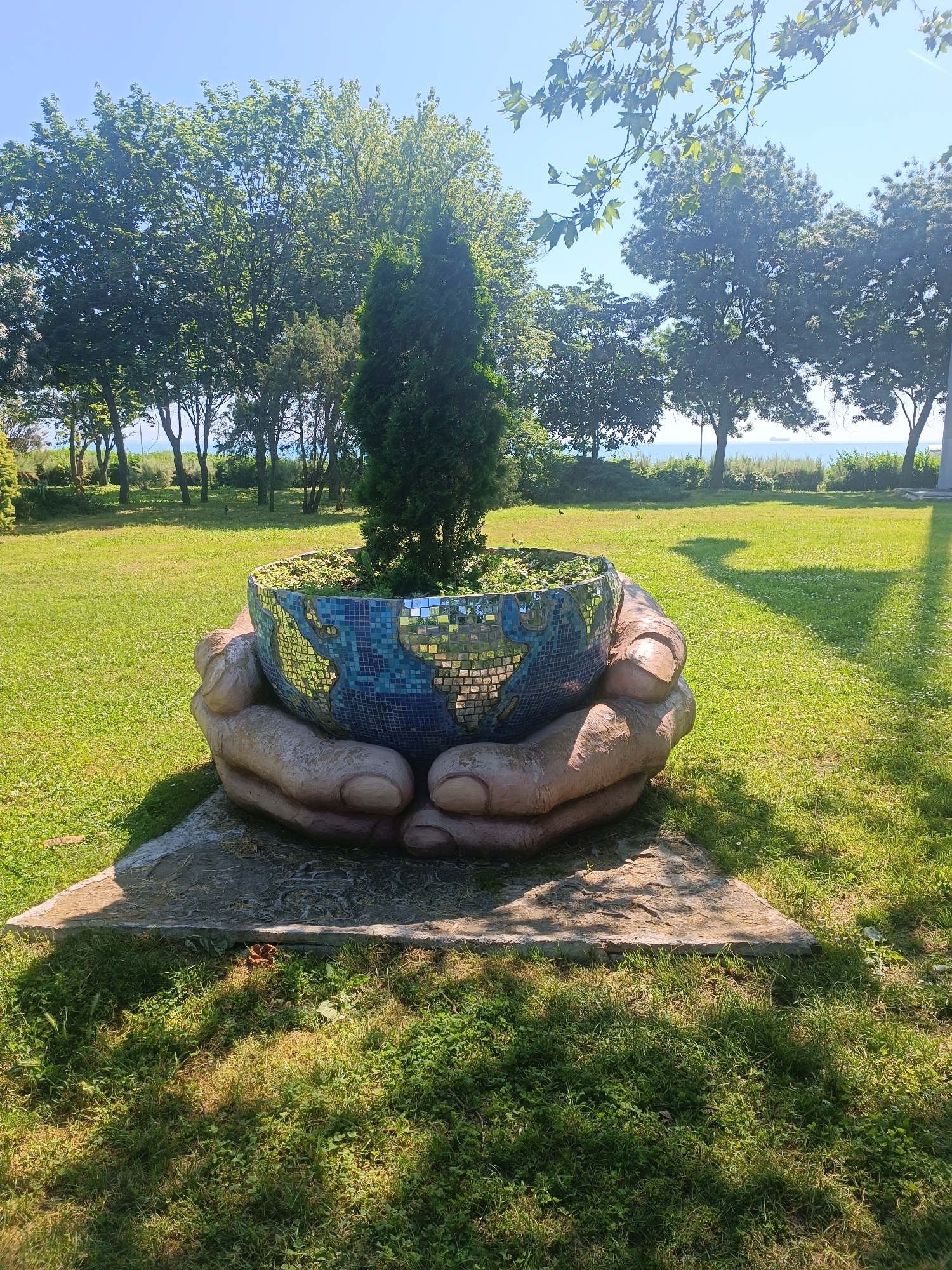 The image depicts a sculpture of two cupped hands holding a mosaic globe planter outdoors. The globe is decorated with blue and white tiles, representing water and land, and is filled with soil and a small evergreen tree. The sculpture is situated on a concrete base, which rests on a patch of grass. Behind the sculpture, a lush green lawn stretches towards a line of trees, and beyond that, the sea is visible under a clear blue sky. The scene is bathed in sunlight, casting shadows on the grass, suggesting a sunny day. The location is the Sea Garden in Burgas, Bulgaria. There are no people visible in the image.