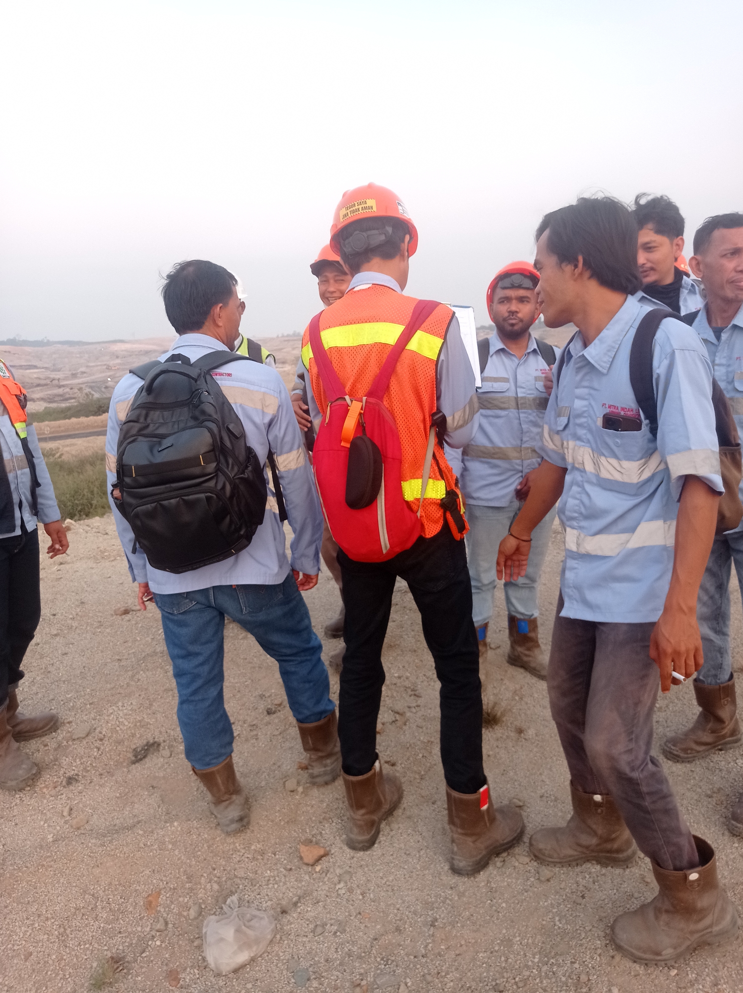 The image shows a group of men, likely construction or mine workers, gathered outdoors on a dusty, uneven surface. The environment appears to be an open, arid landscape with low-lying hills or spoil piles in the background. The sky is overcast or hazy.
Several men are wearing light blue long-sleeved shirts with reflective stripes, dark pants, and brown work boots. Some are also wearing hard hats, with one man in the foreground wearing an orange hard hat with text that reads "TEGUR SAYA JIKA TIDAK AMAN" (Which translates to "Warn me if it's unsafe"). Another man is wearing a bright orange high-visibility vest over his shirt. Backpacks are visible on at least two individuals. One man in the center is holding a clipboard or document.
The men appear to be engaged in a discussion or briefing. Their postures suggest a work-related meeting. One man in the foreground is holding a lit cigarette. The general atmosphere is utilitarian and industrial.
The location is identified as being in the city of Wonomulyo, Indonesia, which is a recognized area for mining activities.