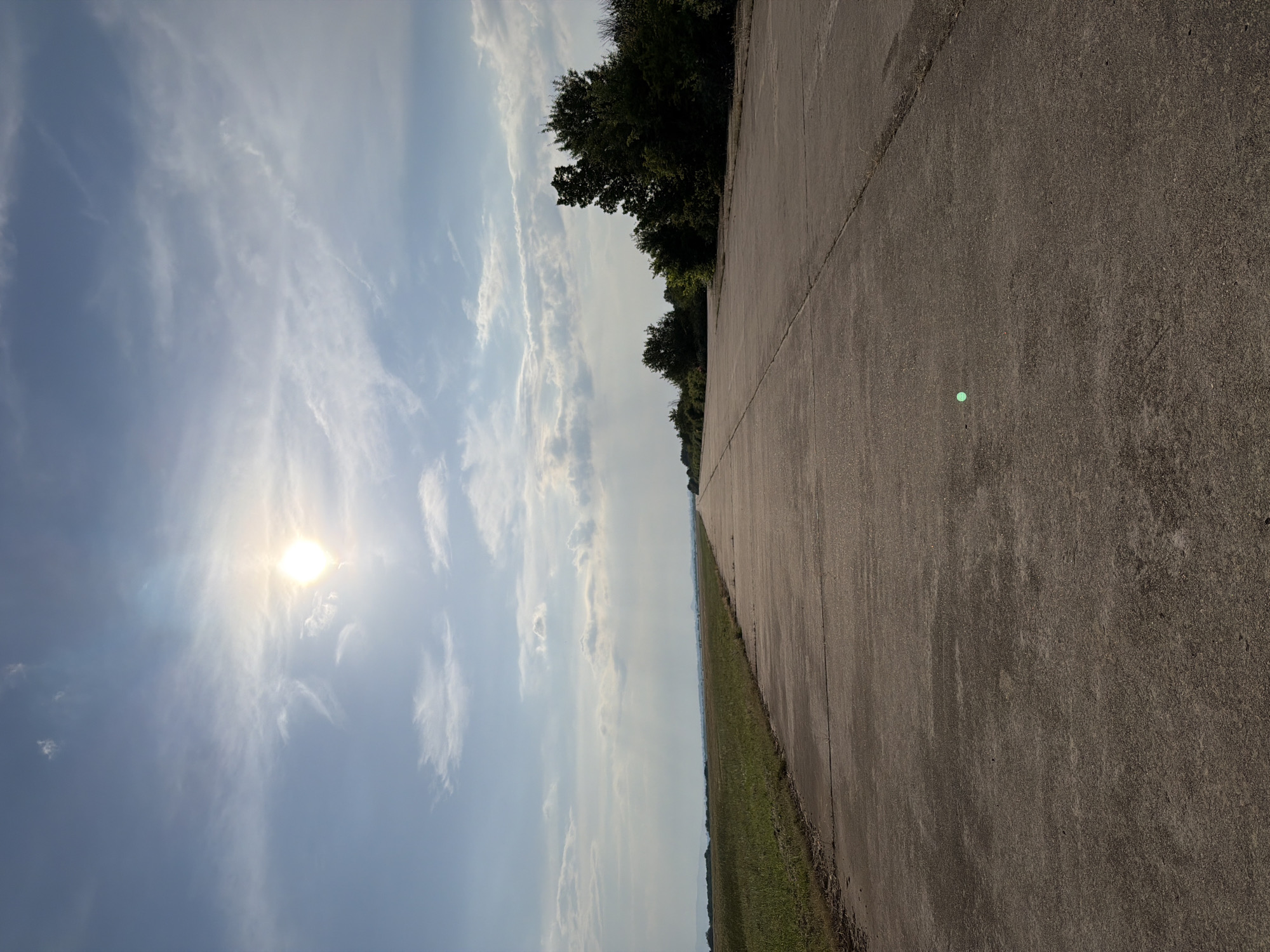 The scene depicts a long, straight concrete road, possibly a disused runway or service road, under a partly cloudy sky in Bogdanitsa, Bulgaria. The road occupies the lower half of the frame, extending from the foreground to a distant vanishing point on the horizon. The concrete surface is gray, weathered, and shows visible seams and minor cracks. In the lower-middle foreground, a distinct small, bright green circular object, consistent with a lens flare artifact, is present on the road surface.
To the left of the road, a wide expanse of green, uncultivated grass or field stretches towards a hazy horizon where faint, distant landforms or hills are visible. To the right of the road, a dense treeline composed of various deciduous trees and bushes runs parallel to the concrete, casting soft shadows towards the left.
The sky is light blue with scattered white clouds, including wispy cirrus and more substantial cumulus formations. The sun is prominently visible in the upper left quadrant of the sky as a bright, overexposed disk, creating a noticeable lens flare and illuminating the scene from what appears to be a late morning or late afternoon angle. No people, animals, or vehicles are present in the image.