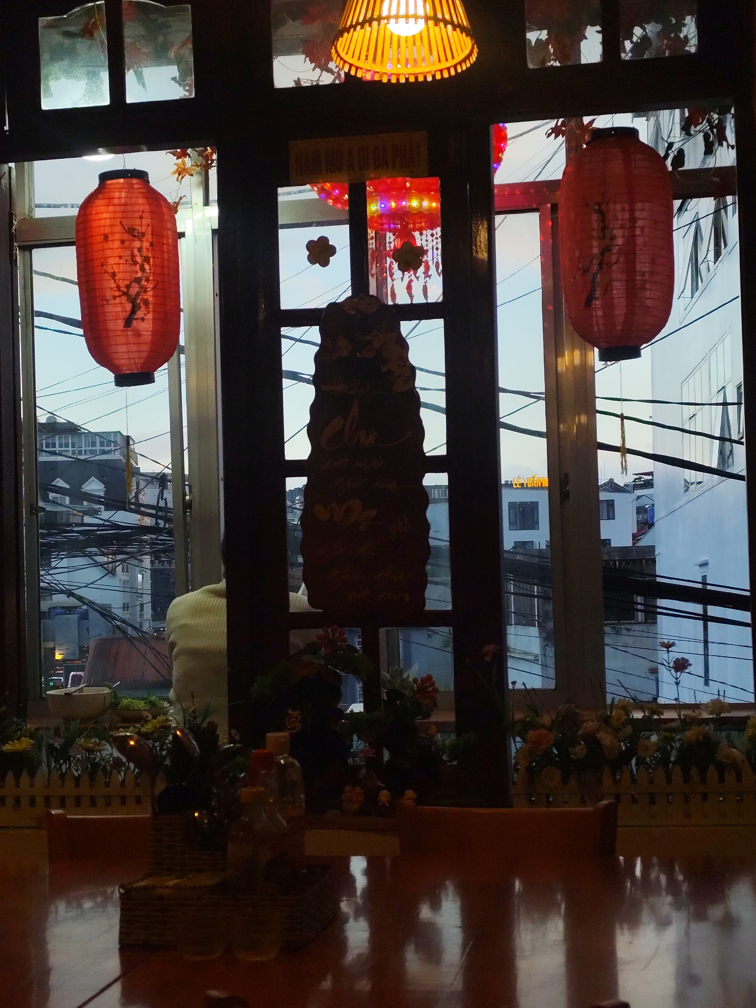 The image depicts an interior scene looking out a window onto a city street, likely in Da Lat, Vietnam, given the Vietnamese signage. In the foreground, a reflective wooden table is visible, with a woven basket containing bottles and possibly some condiments. Beyond the table, a white picket fence-like structure separates the indoor space from the outdoor view.
Through the window, two large red, cylindrical lanterns with black tops and bottoms are suspended. The lanterns appear to be made of paper and are decorated with what looks like a branch with blossoms. A third, smaller, multi-colored lantern hangs further back, adorned with small dangling decorative elements. Above the lanterns, a wooden lampshade with a visible bulb is partially visible, casting a warm light.
The view outside the window reveals a dense urban environment. Buildings of various sizes and colors, primarily white and grey, are clustered together. Numerous electrical wires crisscross the scene, a common sight in Vietnamese cities. A sign with Vietnamese characters, "NAM HOA A O DA PHAT," is visible on the exterior wall of one building. The sky is overcast, suggesting it may be evening or a cloudy day.
A person, seen from behind, is seated at the table, wearing a light-colored, possibly yellow, sweater. They are looking out the window. The overall atmosphere suggests a restaurant or cafe setting, decorated with traditional Vietnamese elements like lanterns.