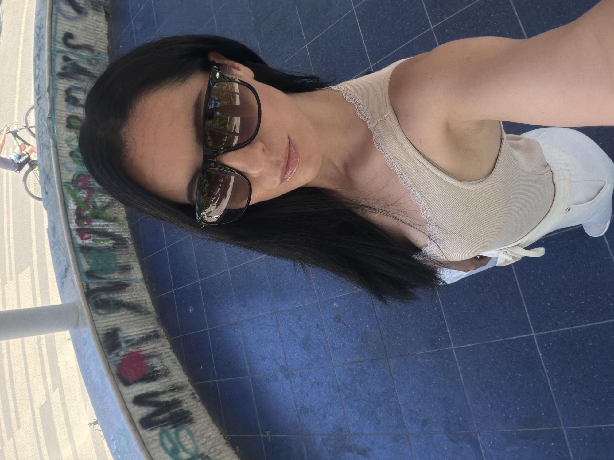A woman with long, dark hair and sunglasses is taking a selfie outdoors. She is wearing a beige ribbed tank top with lace trim and white pants. She is standing on a blue tiled ground. Behind her, a curved concrete wall is covered in graffiti. In the background, there is a person riding a bicycle. The scene appears to be an urban public space, possibly a park or plaza. The specific location in the city of Pleven, Bulgaria, is not identifiable from the visual information provided.