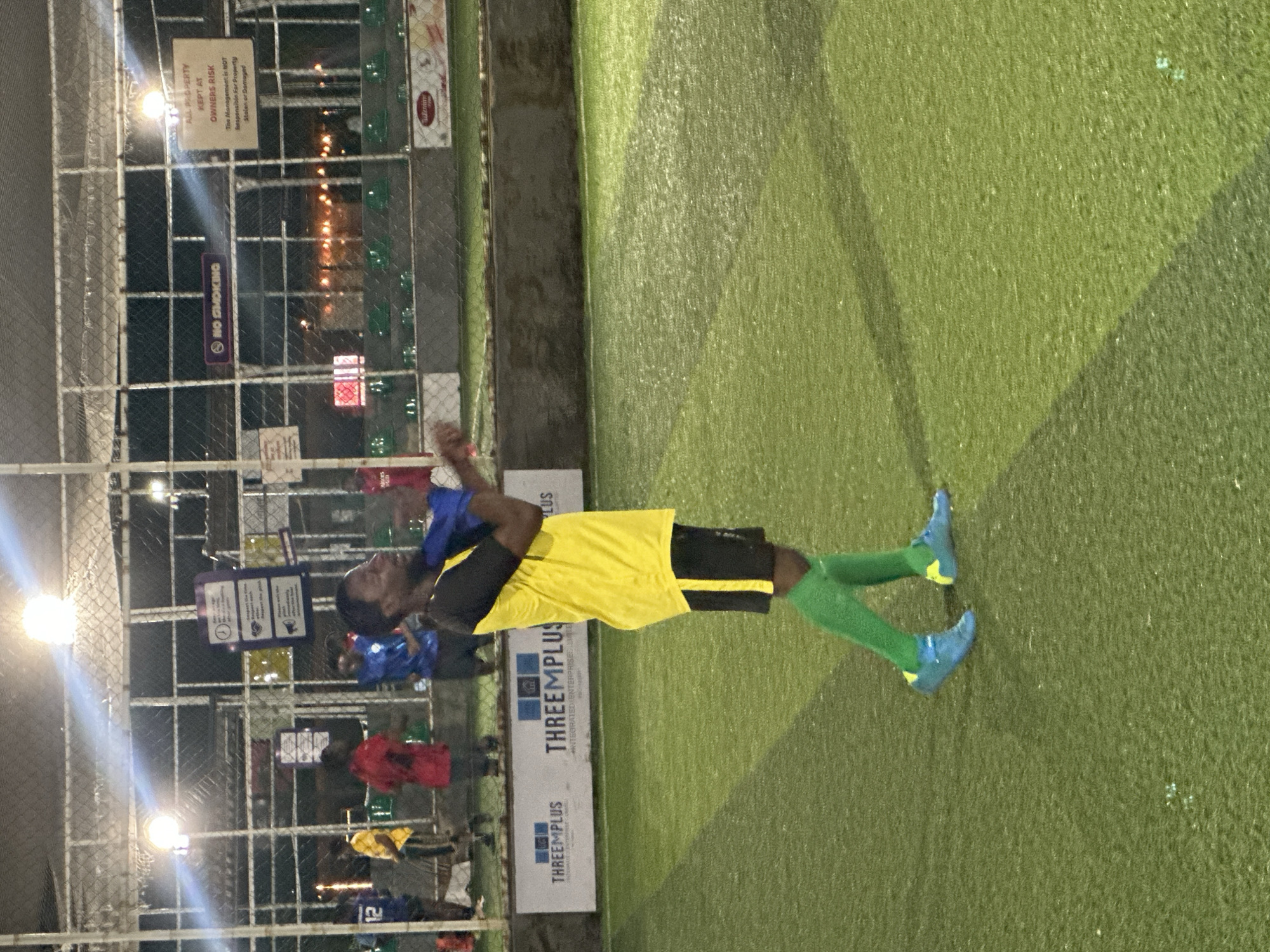 An outdoor artificial turf soccer field in Abuja, Nigeria, is illuminated by overhead lights at night. A Black male soccer player, wearing a yellow and black jersey, black shorts, green socks, and blue cleats, is standing in the middle of the field, facing to the right. He is performing a stretching exercise, holding his arms up and behind his head. Other players in red, blue, and yellow jerseys are visible in the background, as well as spectators sitting on green stadium seats. Fencing surrounds the field, with several signs posted, including a "NO SMOKING" sign and a sign stating "ALL PROPERTY KEPT AT OWNER'S RISK." The Threemplus logo is visible on a banner in the foreground.