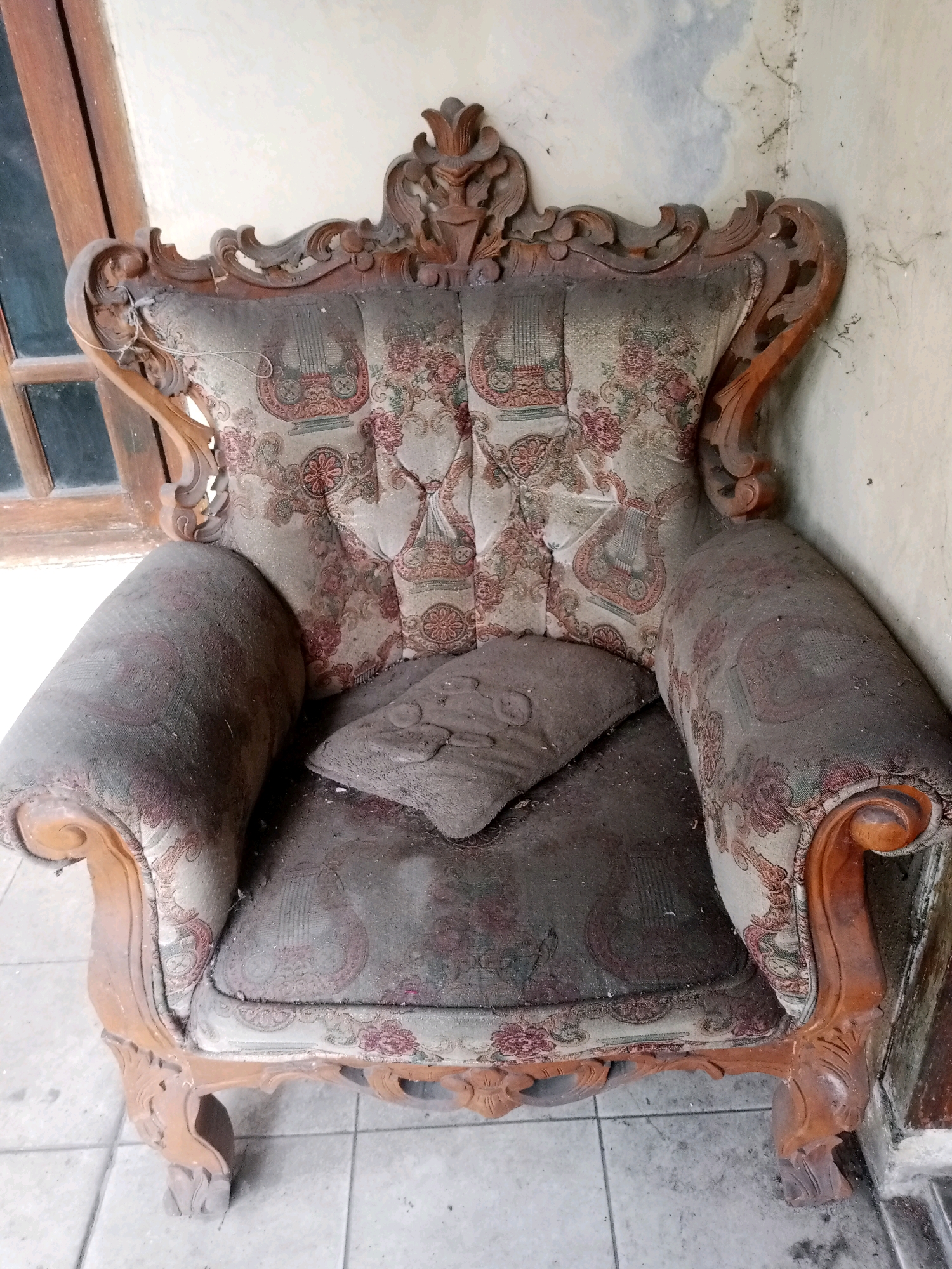 An antique armchair with ornate wooden carvings and floral patterned upholstery, covered in dust, is positioned on a tiled floor next to a wall and a wooden-framed window. A small, similarly dusty cushion rests on the seat. The scene appears to be within an indoor space, possibly a residence, in Balaraja, Indonesia, suggesting a state of disuse or neglect.