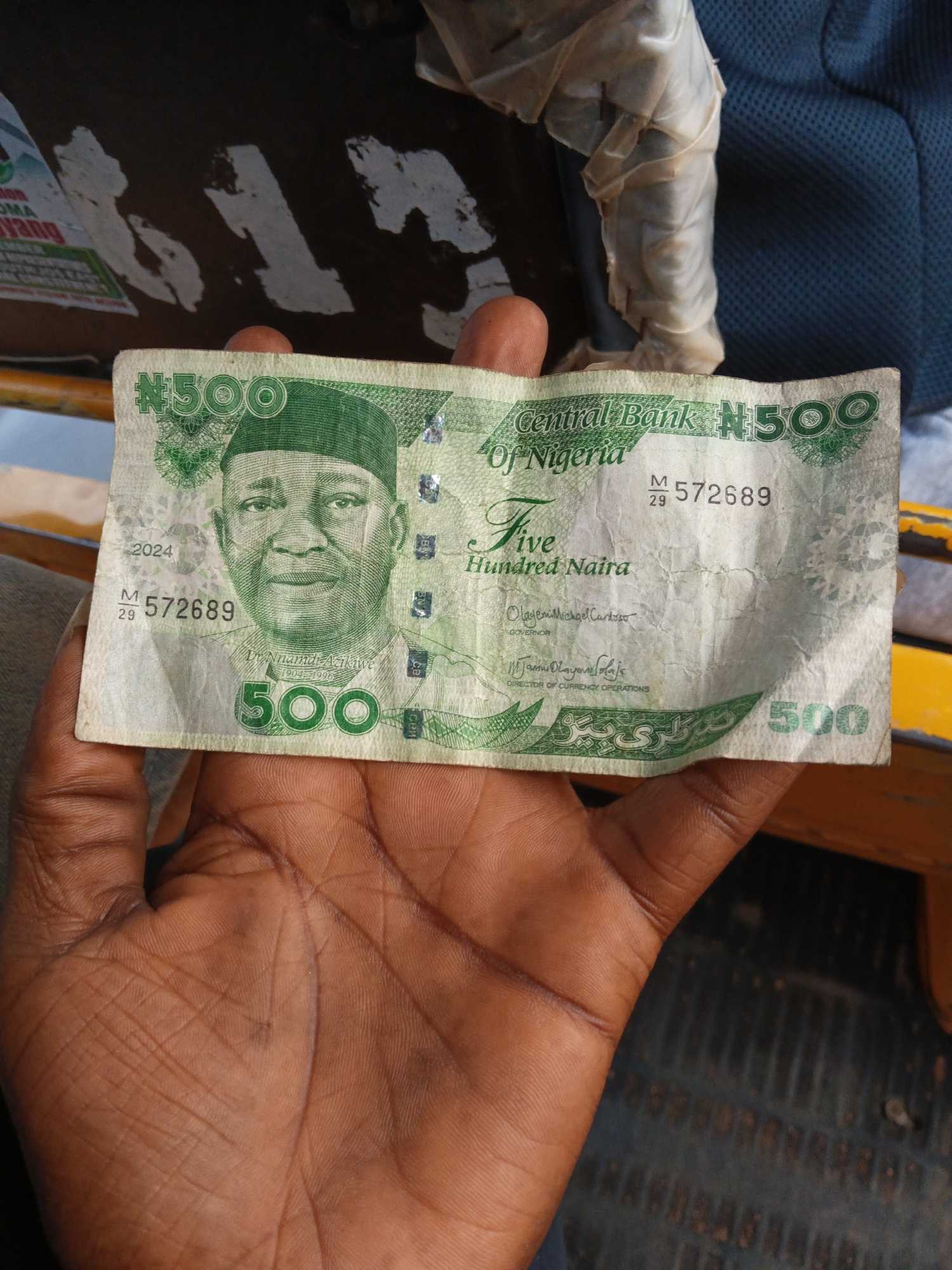 A Nigerian 500 Naira note is held by a person's hand. The banknote is green and features the portrait of Nnamdi Azikiwe. The text "Central Bank Of Nigeria" and "Five Hundred Naira" are visible. The serial number "M 29 572689" is printed on the note. The background is blurred and appears to be outdoors, possibly in a vehicle, with the presence of a blue textured surface and yellow elements.