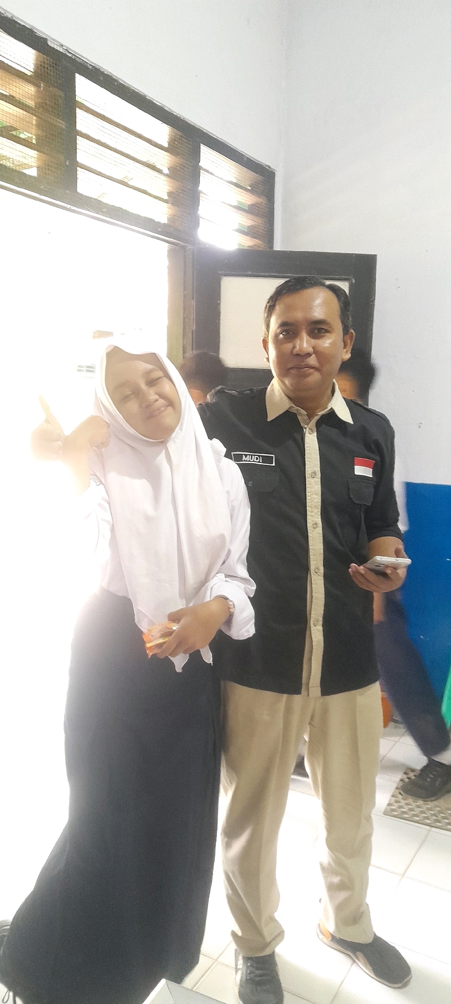 A medium shot captures two individuals indoors, with a window providing bright natural light on the left side. A young female wearing a white hijab and school uniform, including a black skirt, stands on the left, holding a snack in her right hand and giving a thumbs-up with her left. To her right, a man in a black collared shirt with light-colored trim and khaki pants stands, holding a mobile phone in his right hand. A name tag on his shirt reads "MUDI," and a small Indonesian flag patch is visible on his right chest pocket. In the background, another person is partially visible, along with a door and a blue cabinet. The floor appears to be tiled. The overall impression is a casual moment captured within what seems to be a school or administrative building in Tahunan, Indonesia.