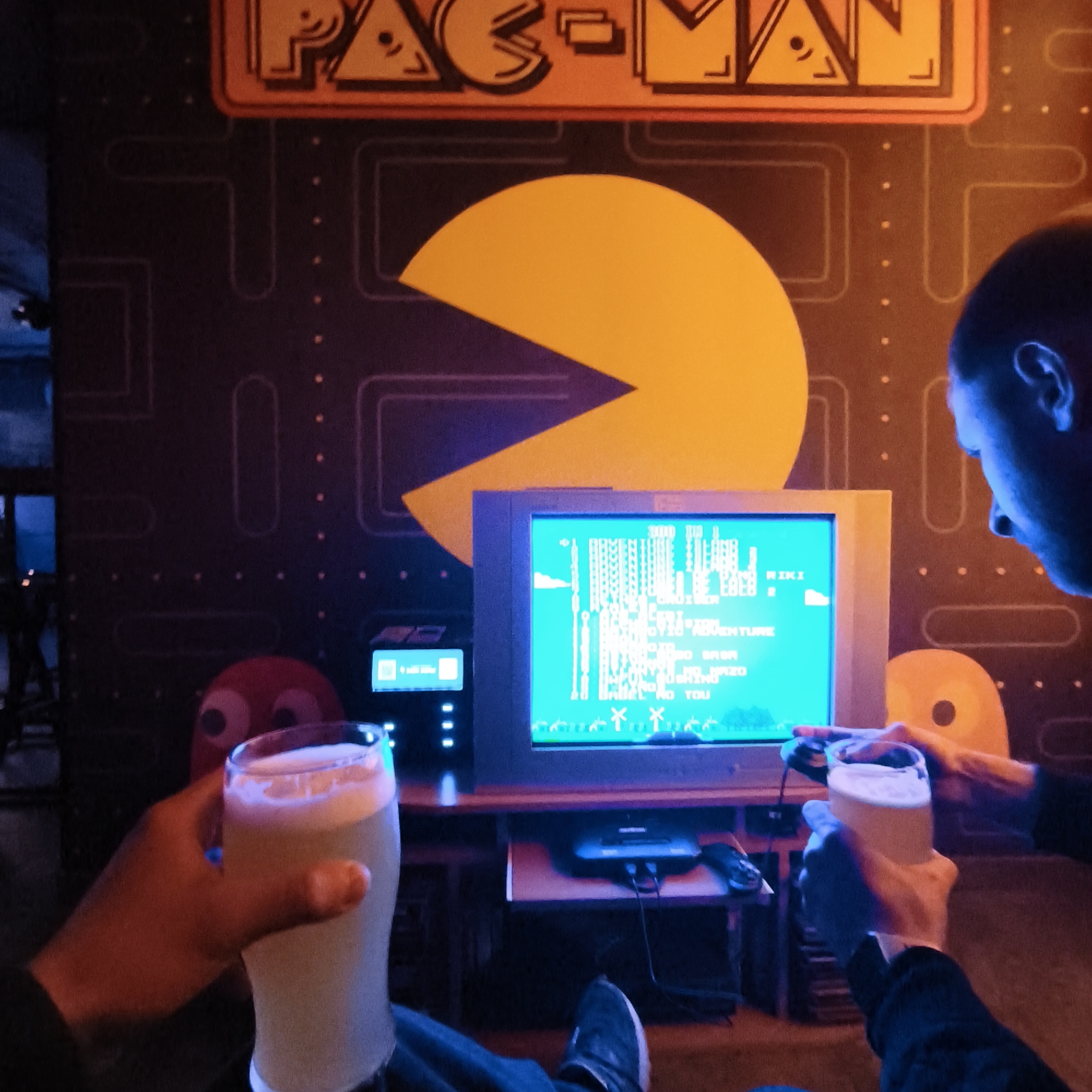 Two people are sitting in an arcade-style room, possibly a bar or entertainment venue in Hrodna, Belarus, in front of a CRT television screen displaying an 8-bit style game. The background wall features a Pac-Man design with a large yellow Pac-Man graphic and a Pac-Man logo above. One person is visible from the side profile, looking at the screen. Each person is holding a pint of beer in their hand. A Sega Genesis console is located under the TV. A red Pac-Man ghost plush toy is visible near the edge of the screen, and a smaller screen displaying game options is also visible next to the CRT TV.