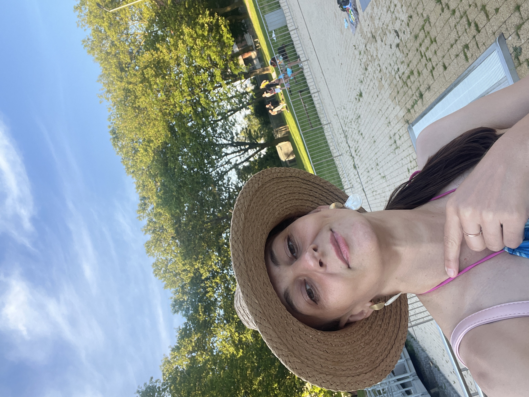 A woman in a straw hat and bikini top takes a selfie outdoors near a body of water. She is wearing large, white earrings and a ring on her right hand. In the background, people are visible near trees and a fenced-off area, possibly a swimming pool. The sky is blue with wispy clouds.