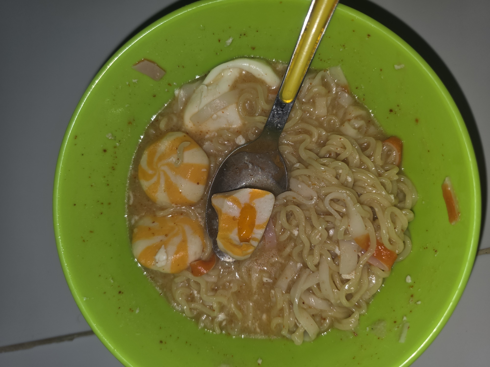 A top-down, close-up shot shows a green plastic bowl containing noodles, broth, egg slices, and orange and white spiral-patterned fish balls. A metal spoon with a yellow handle rests within the bowl. The bowl sits on a light grey tiled surface. The scene appears to be indoors, illuminated by artificial light. The image was taken in Pintomakmur, Indonesia.