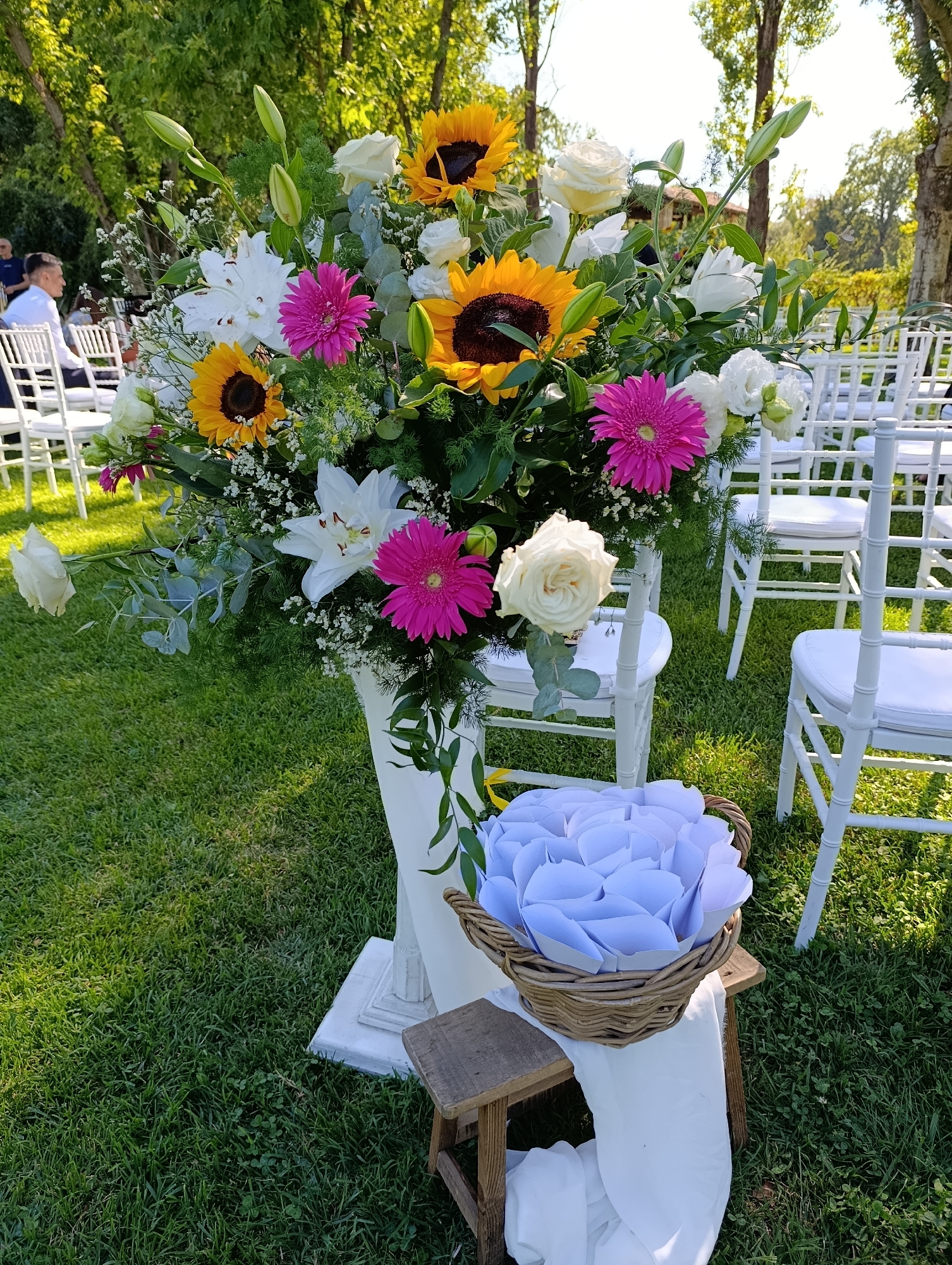 A tall floral arrangement featuring sunflowers, lilies, roses, and pink gerbera daisies stands in a white pedestal on a grassy lawn. The arrangement is adorned with greenery and baby's breath. To the right of the floral arrangement, a woven basket filled with white paper cones sits on a small wooden stool, draped with white fabric. In the background, rows of white chairs are set up on the lawn, suggesting an outdoor event. A person in a white shirt is visible in the far left background, seated. The scene is illuminated by bright, natural sunlight, casting shadows on the grass and highlighting the vibrant colors of the flowers. The environment appears to be a park or garden setting.