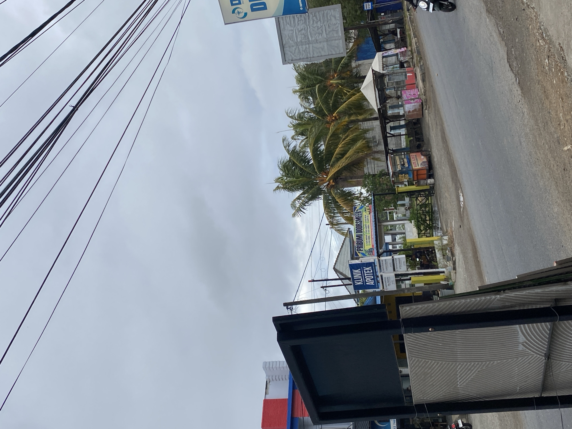 The image depicts a street scene in Payatieng, Indonesia, on an overcast day. In the foreground, a large black frame with a textured gray panel is positioned on the left. The street itself is paved with asphalt, and a motorcyclist is visible on the right side of the road, heading away from the viewer.
Along the street, several small businesses are apparent. A blue and white sign reads "KLINIK APOTEK" (Clinic Pharmacy). Next to it, another sign advertises "PRIBUMI DOORSMEER" which appears to be a car wash or detailing service, with smaller text listing services like "CUCI MOTOR" (motorcycle wash) and "CUCI MOBIL" (car wash). Further down the road, several food stalls with awnings are set up. One stall has a sign that says "SATE PADANG" and "SATE AYU," indicating it sells Sate Padang and another type of Sate. Another stall has pink and white branding.
Palm trees are visible behind the stalls, their fronds swaying slightly. In the background, buildings with red and white facades, as well as a light gray building with what looks like corrugated metal siding, are present. A white sign with a blue logo and the letter "D" is also visible. Power lines crisscross the sky, a common sight in urban and semi-urban Indonesian settings. The overall atmosphere is that of a typical commercial street in Indonesia.