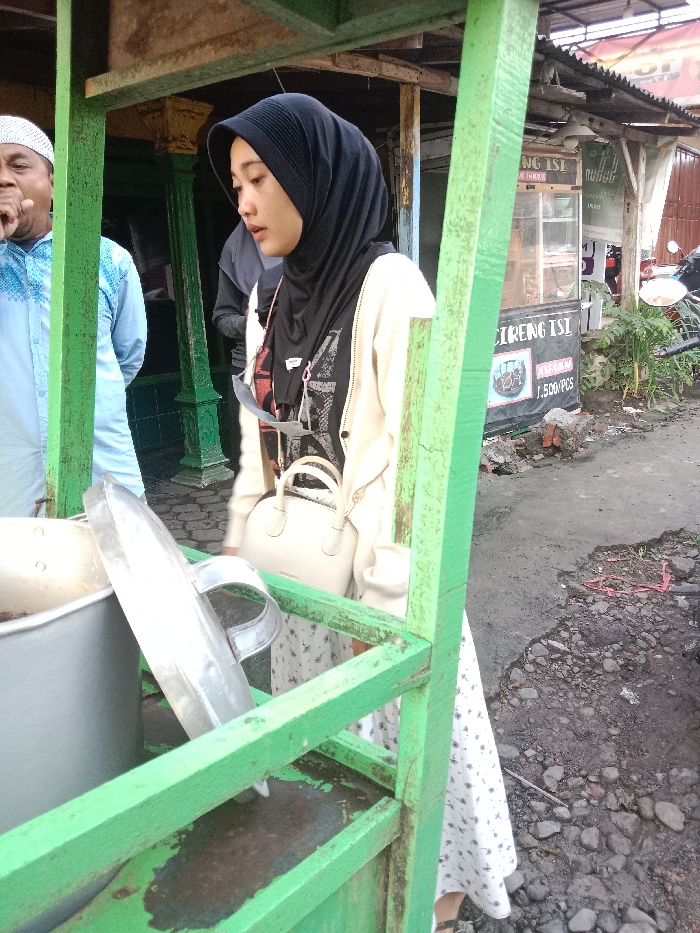 A woman in a black hijab and cream cardigan stands near a food stall constructed with green wooden frames. A large metal pot and lid are visible on the stall. A man wearing a white skullcap and blue patterned shirt stands to the left of the woman. Behind her, a grey building can be seen, with signage reading "CIRENG ISI." A motorbike is parked in the background to the right. The ground is paved with grey concrete.