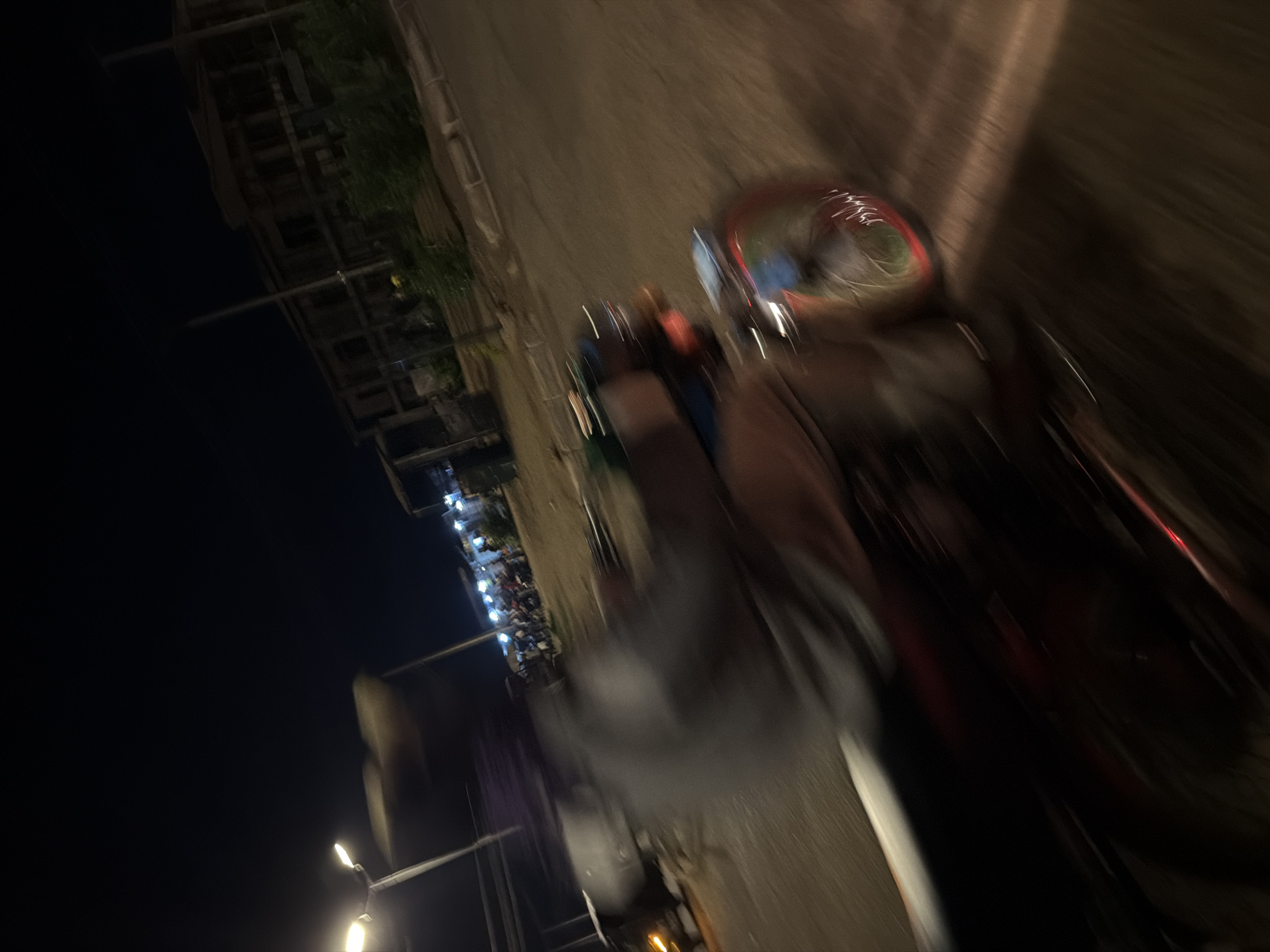 This is a low-light, motion-blurred image captured at night on a paved road in Bukuru, Nigeria. The primary subject is a person riding a motorcycle, depicted as a streak of motion. The rider is wearing a light-colored shirt. The motorcycle has a red frame and a colorful wheel rim. In the background, there are streetlights illuminating a collection of stalls or shops, with several other motorcycles and people visible, suggesting a marketplace or gathering area. A multi-story building, possibly under construction, is also visible in the distance. The overall scene conveys a sense of movement and activity at night.