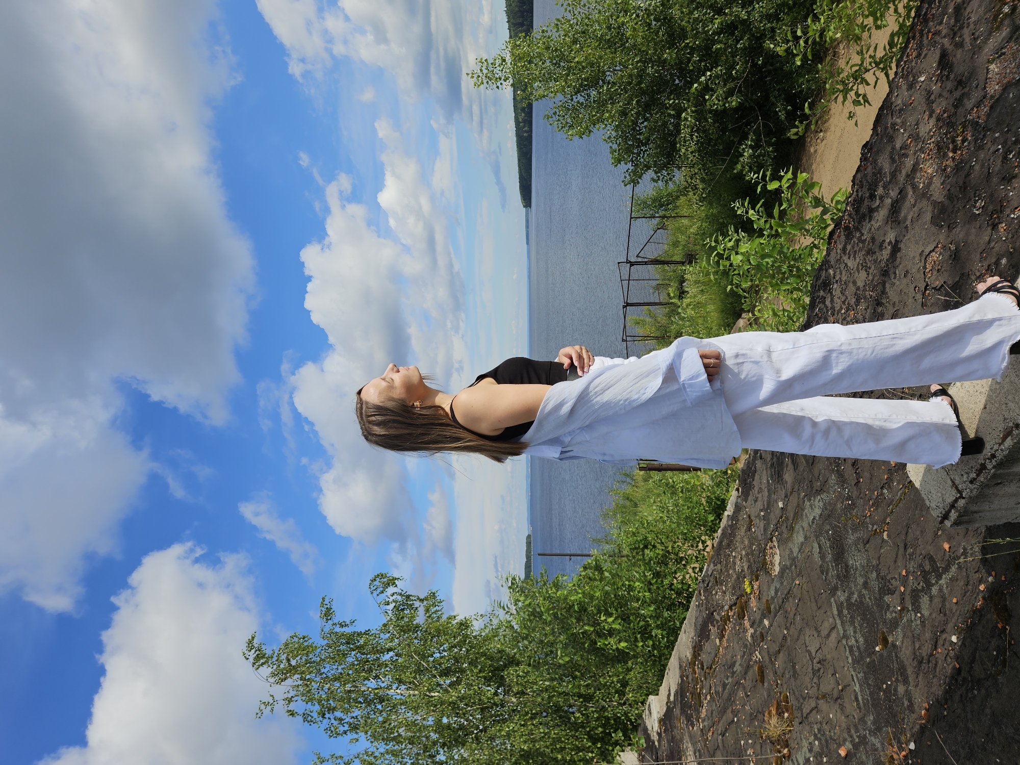 A young woman stands on a weathered concrete structure overlooking a wide body of water, likely the Kama River, near Chaikovsky, Russia. She is dressed in a black tank top and white, wide-leg pants, with a light blue open shirt draped over her shoulders. Her face is turned upwards, eyes closed, as if basking in the sun. The background features a vast expanse of blue sky dotted with fluffy white clouds, lush green trees, and the distant shoreline. The concrete platform appears to be an old, possibly abandoned structure, with visible cracks and debris. The overall scene suggests a peaceful, contemplative moment amidst a natural landscape.