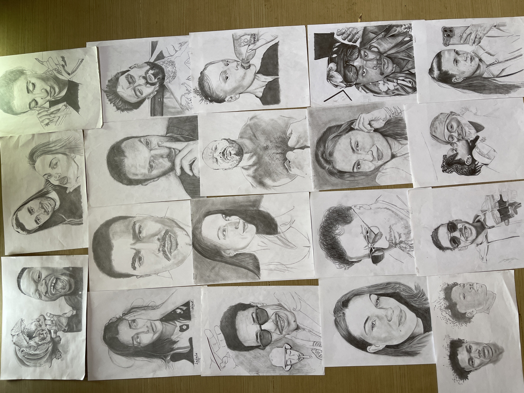 The image displays a collection of twenty-four individual pencil sketches, predominantly portraits, arranged on a light brown, wooden surface, likely an indoor table or floor. The artworks, implying a context or origin within Jos, Nigeria, are presented in a loose grid formation, slightly overlapping in some areas.
The sketches are primarily head-and-shoulder or bust portraits of diverse individuals. Subjects include men, women, and a child, varying in apparent age and ethnicity, depicted with a range of expressions from smiling and contemplative to surprised or posed. Noteworthy specific sketches include: a caricature featuring a roaring hippopotamus and a man with an exaggerated open mouth; several individuals wearing sunglasses; a muscular, bare-chested man clenching his fists; a woman drinking from a "NESTLÉ" branded water bottle; a man holding two mugs of beer; and a woman holding a smartphone, appearing to take a selfie. One sketch depicts a child kissing an older man on the cheek, while another shows two different views of the same young man. The artistic style is consistent, characterized by graphite shading to create realistic textures and depth. The white paper sheets on which the sketches are drawn show minor signs of handling, such as slightly curled edges or faint smudges. The overall scene is well-lit, highlighting the monochrome nature of the artwork.