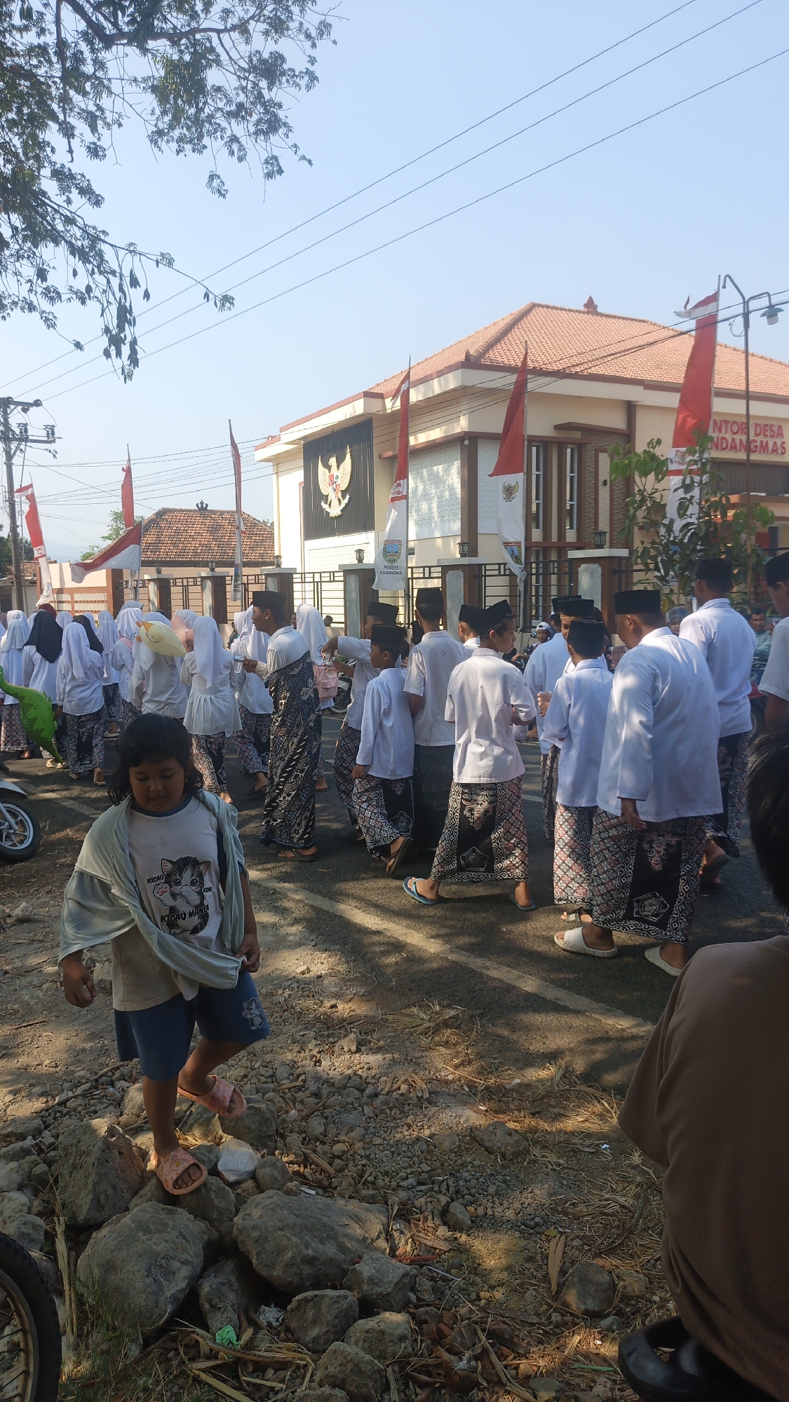 In Kandangmas, Indonesia, a group of children and young people are gathered outside a building, likely a local administrative office, indicated by the text "KANTOR DESA KANDANGMAS" on the facade. The building has a prominent Garuda Pancasila emblem and is adorned with Indonesian flags, suggesting a national or local celebration. Many of the individuals are dressed in traditional Indonesian attire, including white long-sleeved shirts, sarongs, and peci hats. Some are holding decorative items, possibly related to a parade or ceremony. In the foreground, a young child in a light-colored t-shirt and blue shorts stands on a rocky embankment, looking towards the gathering. The scene is set outdoors on a bright, sunny day, with trees and power lines visible in the background.