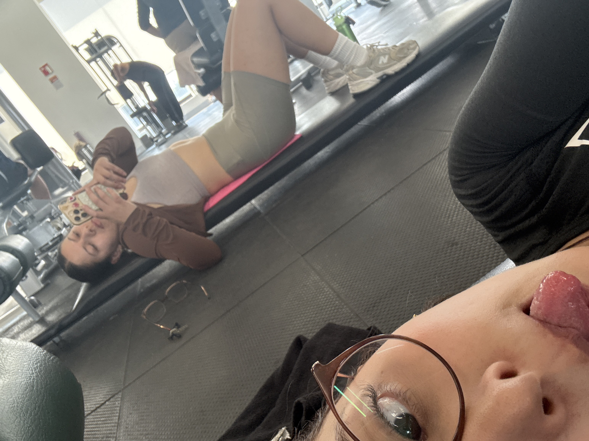 This image captures a casual moment in a gym or fitness center in San Andrés Cholula, Mexico, taken from a low, upward angle, likely a selfie.
In the immediate foreground, part of a person's face is visible, including one eye behind glasses with dark frames, a portion of the nose, and a tongue playfully sticking out to the side. This person is wearing a black top.
In the mid-ground, a second person is lying on their back on a pink exercise mat placed on the dark, textured gym floor. They are wearing a light-colored sports bra or crop top, light olive green cycling shorts, and white socks with light-colored New Balance sneakers, where the letter "N" is visible on the side of the shoe. This individual holds a smartphone with a colorful, patterned case (possibly floral or geometric designs) in their hands, looking down at the screen. Their eyes appear closed or focused on the phone. A pair of brown-rimmed eyeglasses rests on the floor near them.
In the background, other gym-goers are faintly visible. One person is seen bending over, possibly stretching or interacting with a piece of equipment. Various gym machines, including cable and pulley systems, are distributed throughout the brightly lit space, suggesting an indoor daytime setting. The overall atmosphere is relaxed, with individuals engaged in their fitness routines or resting.