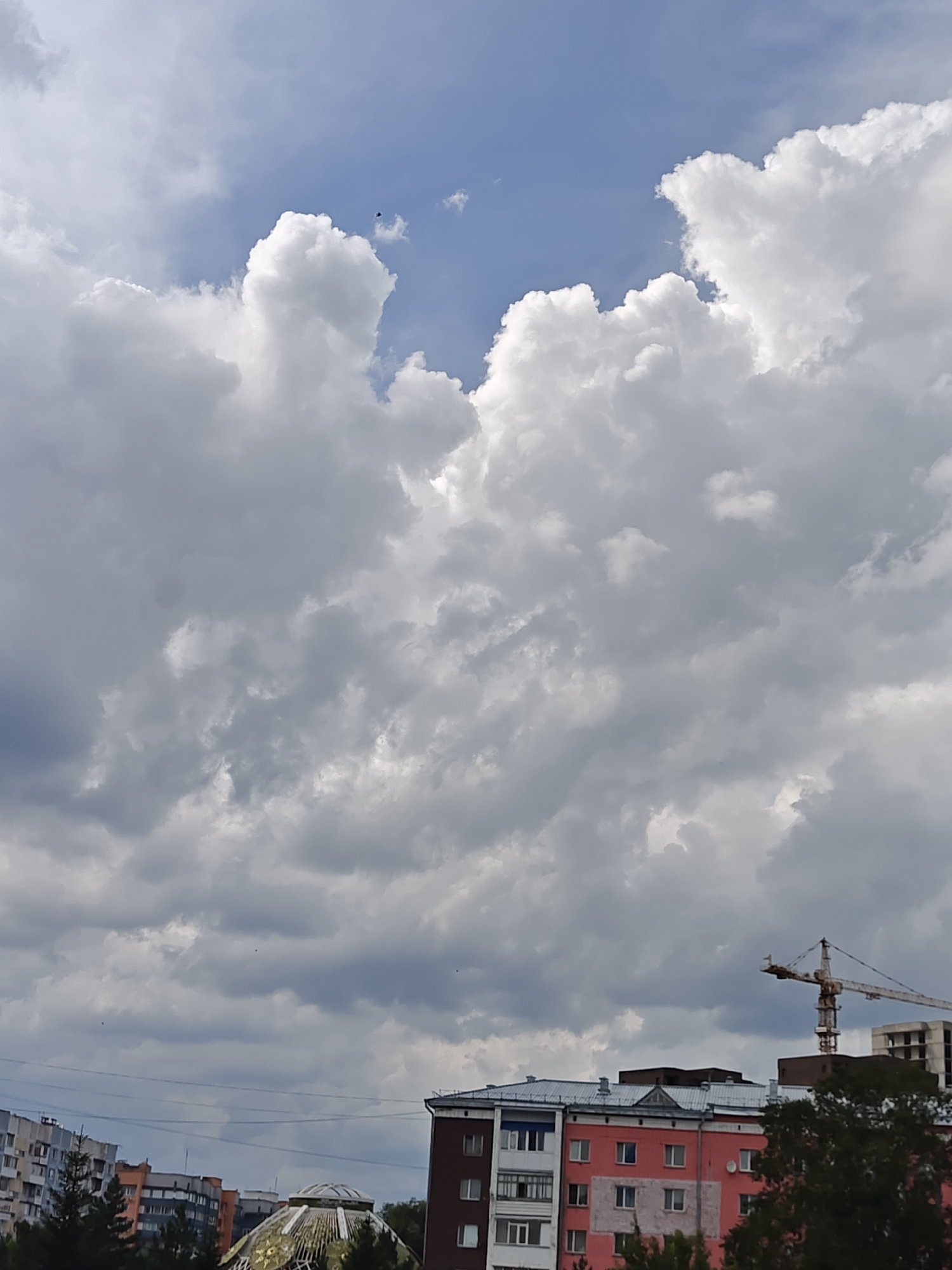 The image captures a view of the sky above a city, likely Petropavl, Kazakhstan, given the context. The sky is a mix of bright blue patches and dense, grey cumulus clouds, suggesting a partially cloudy or overcast day. In the distance, several multi-story buildings are visible, along with trees and a construction crane. One building stands out with a striking pink facade, featuring multiple windows and balconies. A large, domed structure with a metallic, possibly golden, intricate design is also present, partially obscured by trees. The overall scene depicts an urban environment under a dramatic sky.