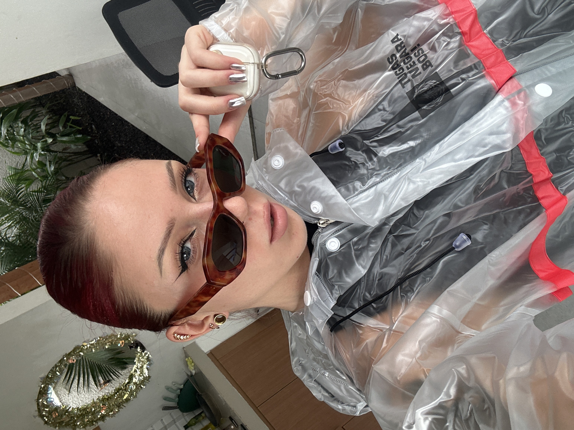 A young woman with reddish-brown hair, styled neatly, looks directly at the camera in this portrait. Her blue eyes are accentuated with winged black eyeliner, and she has full, light pink lips. She wears tortoise-shell cat-eye sunglasses, which are pushed down on her nose, allowing her eyes to be fully visible.
Her attire includes a transparent, clear plastic jacket or raincoat, detailed with white snap buttons down the front and notable red fabric trim along some seams. A black drawstring is visible near the collar, and a dark garment is worn underneath the transparent layer. On the jacket, black text reads "TUGAS NEGARA BOS!".
Her fingernails are painted with a striking silver metallic polish. In her left ear, multiple earrings are visible, including a small gold stud and a decorative ear cuff adorned with sparkling stones. She holds a white wireless earbud case, which has a silver carabiner clip attached to it, positioned near her face.
The background suggests an indoor setting, possibly a modern living space or office. A black mesh office chair is partially visible on the left. Green leafy plants add a touch of nature, and an oval-shaped decorative mirror, embellished with gold tinsel-like trim, reflects a palm leaf, contributing to the stylish ambiance. The scene takes place in North Kuta, Indonesia, during what appears to be daytime.
