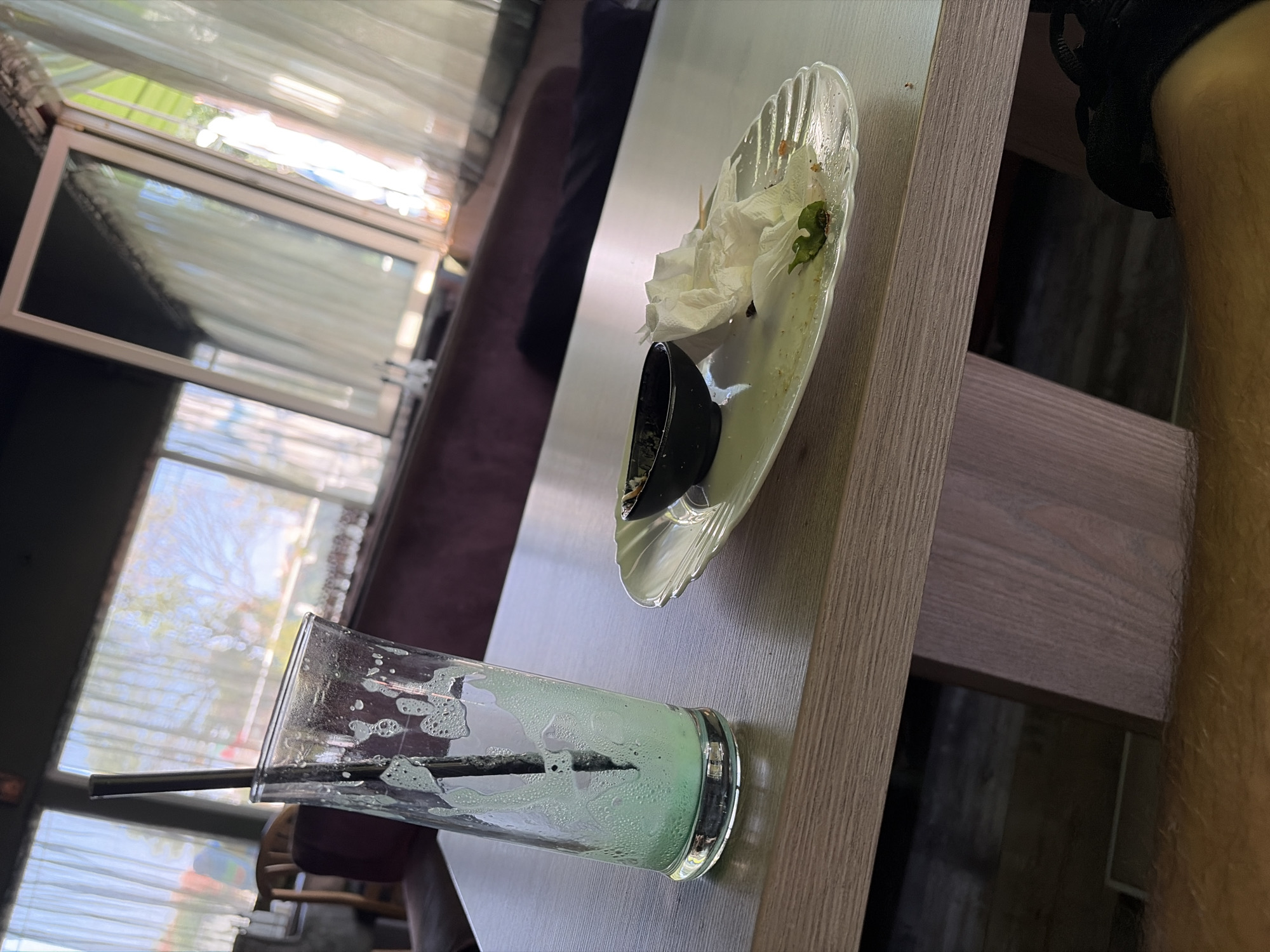 The image depicts a table setting at what appears to be a cafe or restaurant. In the foreground, a tall, clear glass with a straw contains remnants of a green, frothy beverage. Beside it, a white, scalloped-edge plate holds a small black dish, crumpled white napkins, a wooden skewer, and some crumbs. The table itself is light-colored wood with a visible grain. In the background, through large windows with sheer white curtains, an outdoor scene is visible, hinting at a sunny day. The indoor seating area features plush, dark-colored upholstery, likely a booth or sofa. The overall impression is that of a table after a drink and possibly a light meal have been consumed. The location is identified as Elin Pelin, Bulgaria.